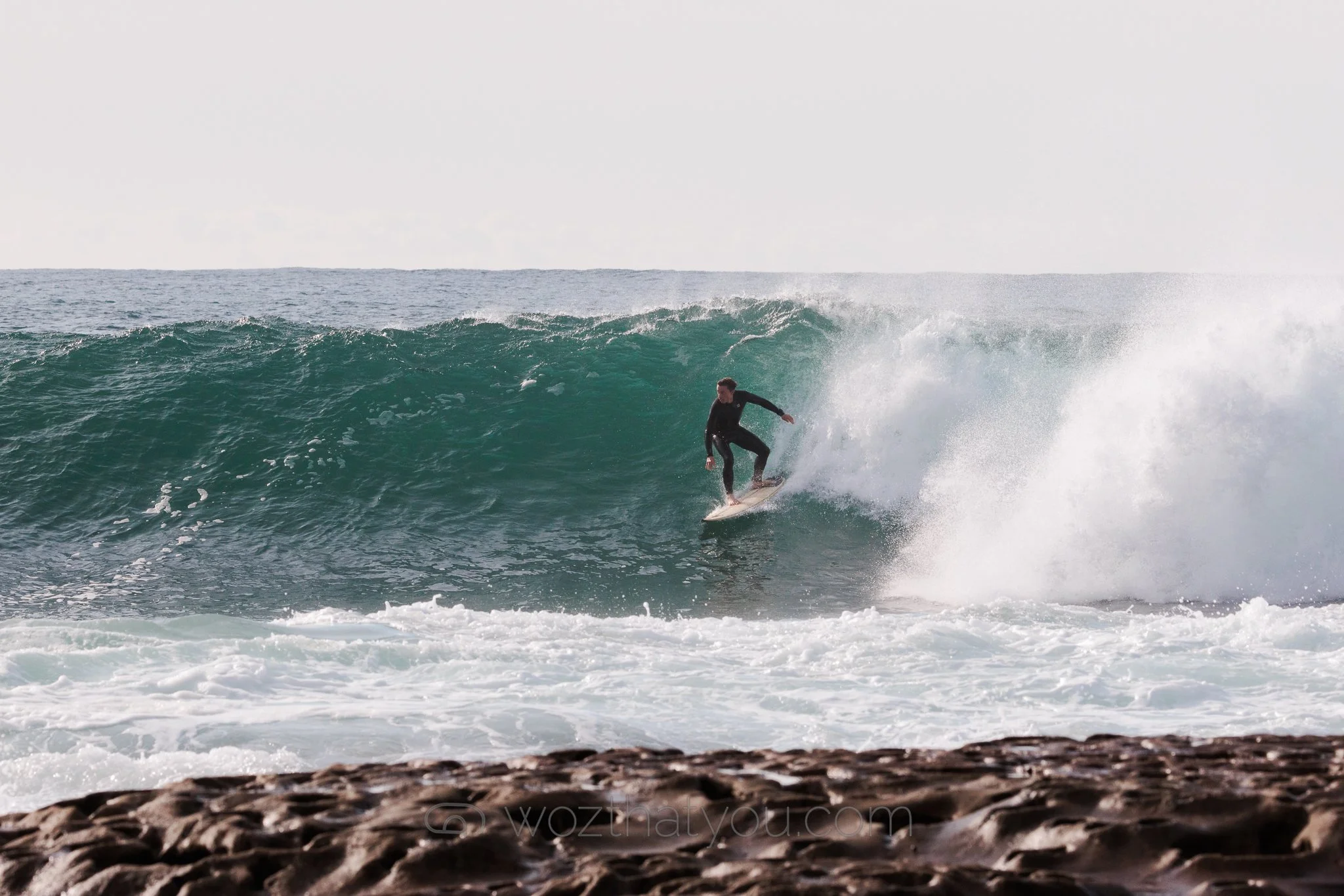 A person in a black wetsuit surfing on a large wave near a rocky shoreline.