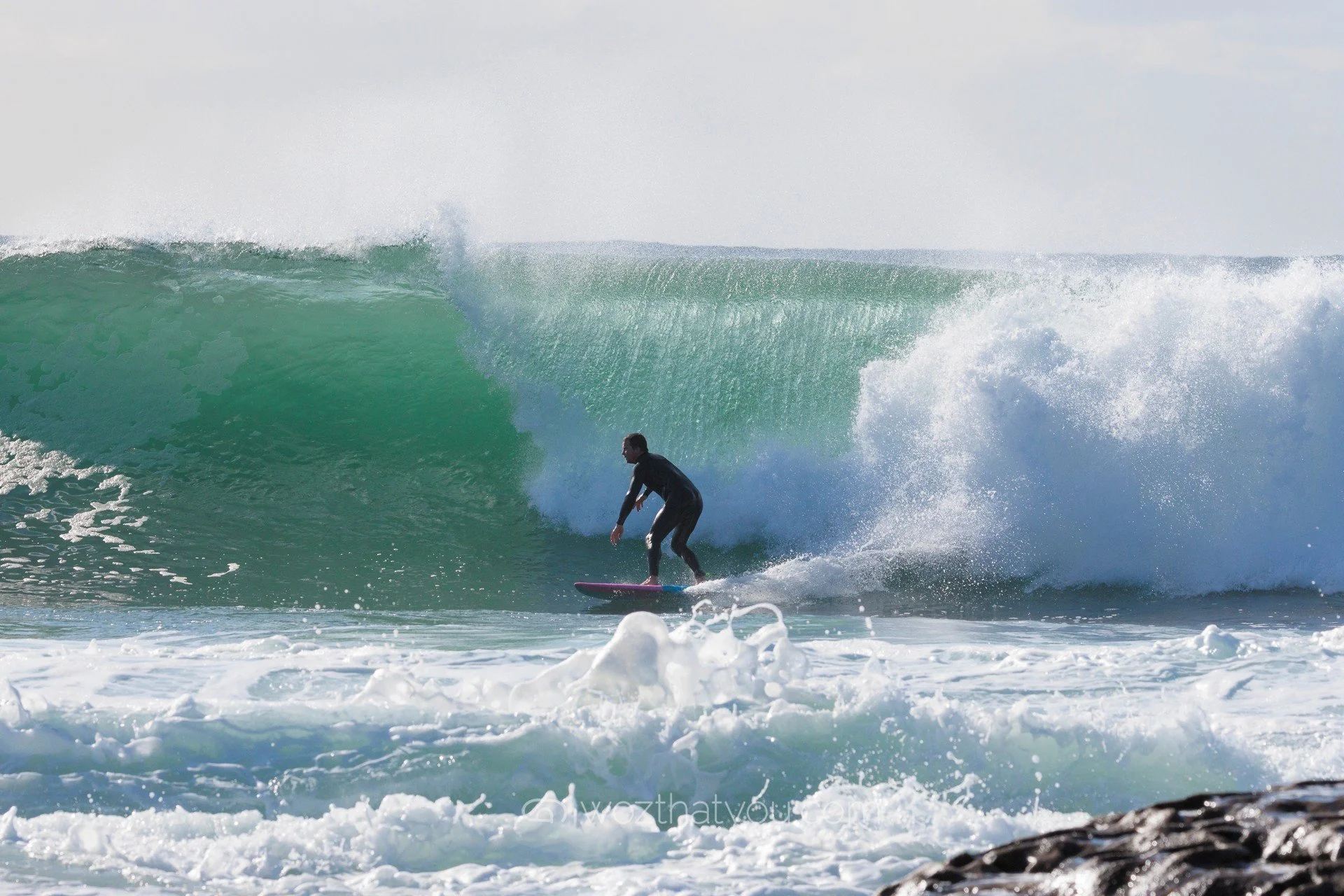 A person wearing a black wetsuit surfing on a pink surfboard on a large ocean wave.