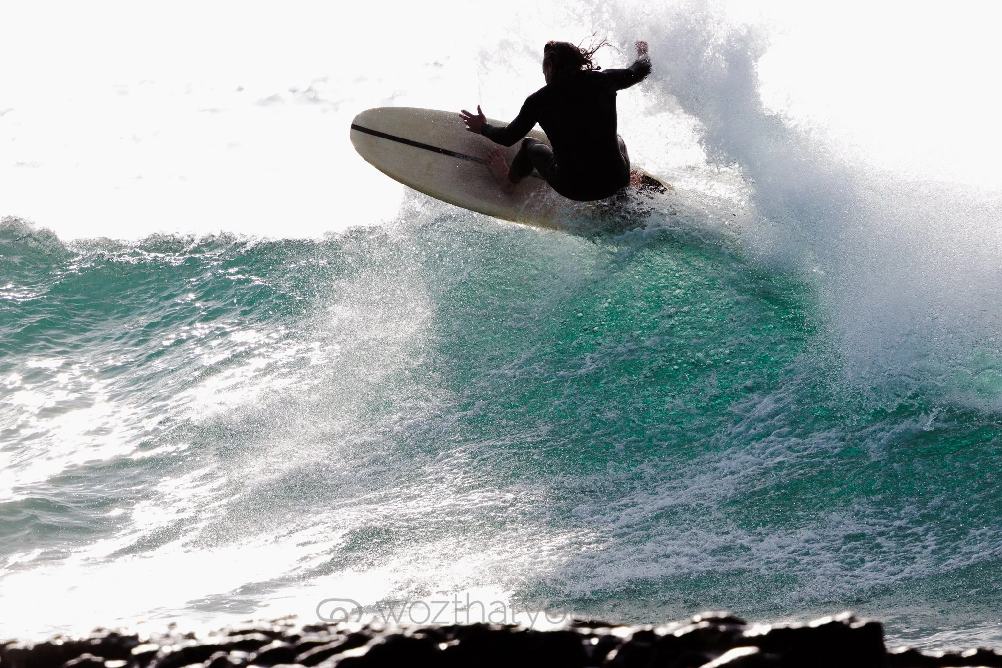 A person surfing on a wave, wearing a wetsuit, with the sun bright in the background.