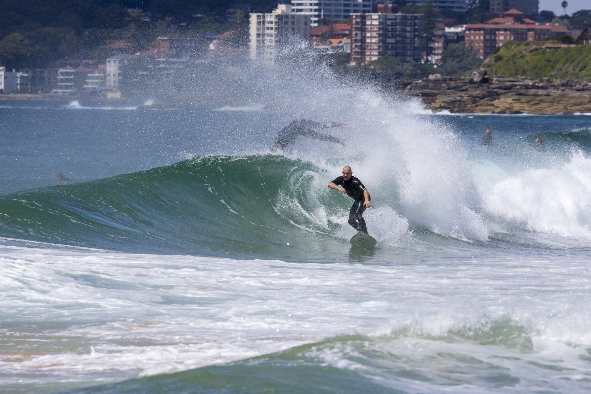 A person surfing on a wave at the beach, with urban buildings in the background.