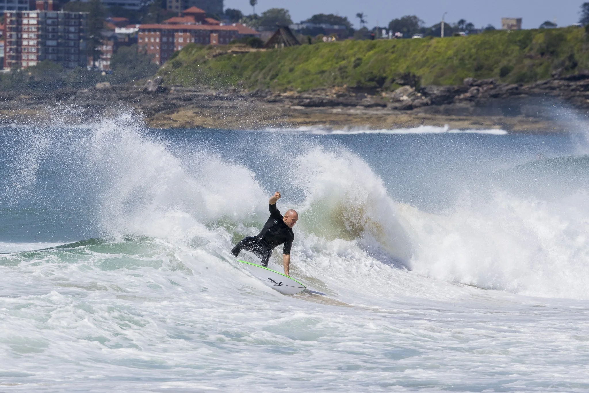 A person surfing on a wave near a rocky shoreline with buildings in the background.