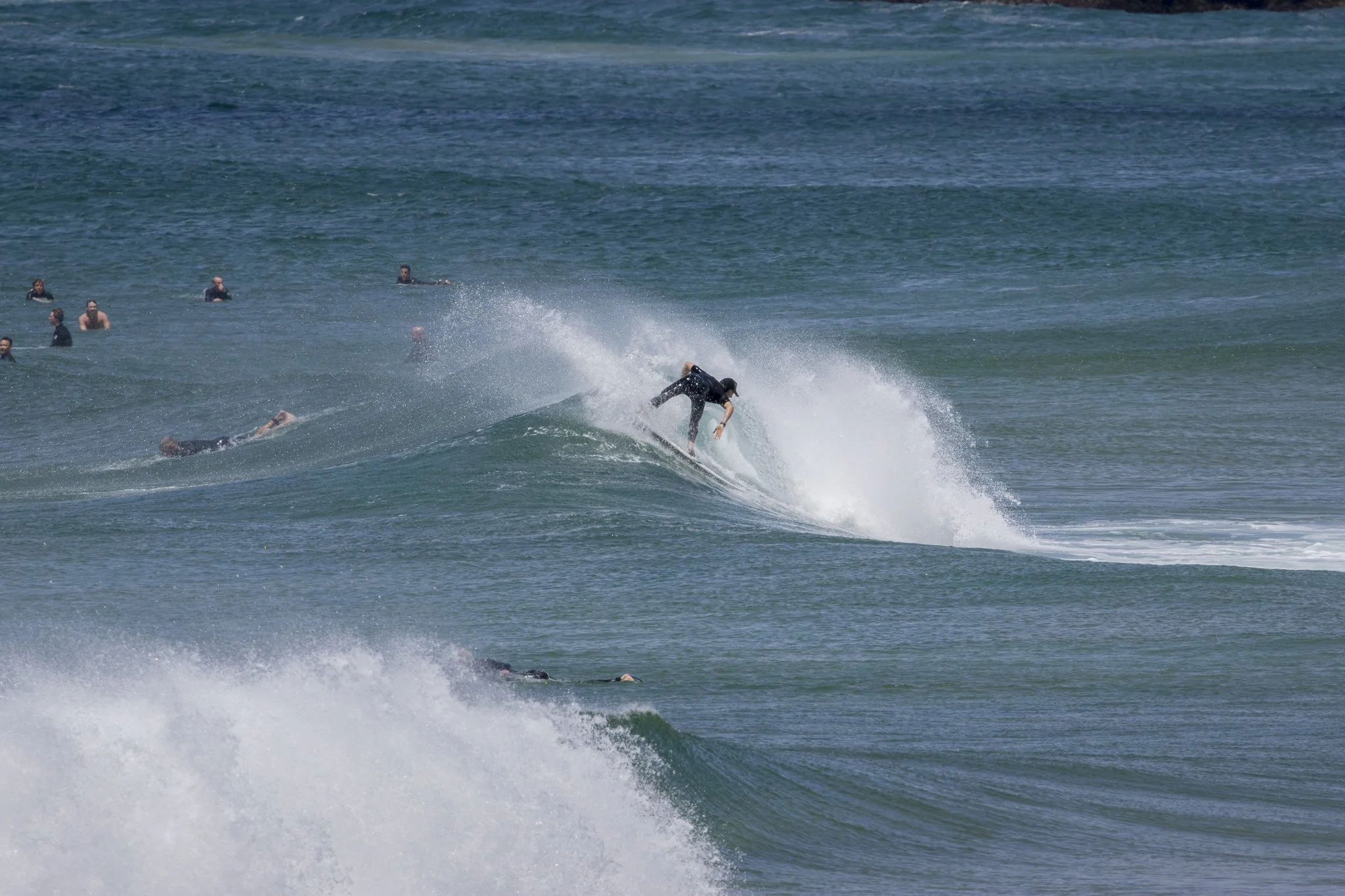Surfer riding a wave while several people swimming are visible in the water in the background.