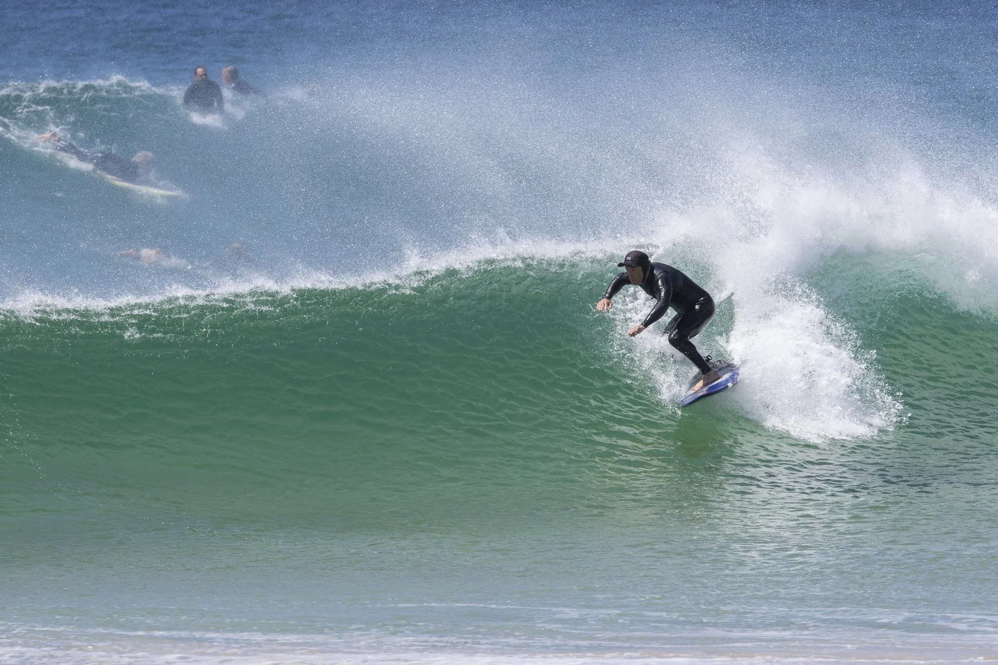 A person wearing a black wetsuit is surfing on a wave at the beach, with other surfers in the background.