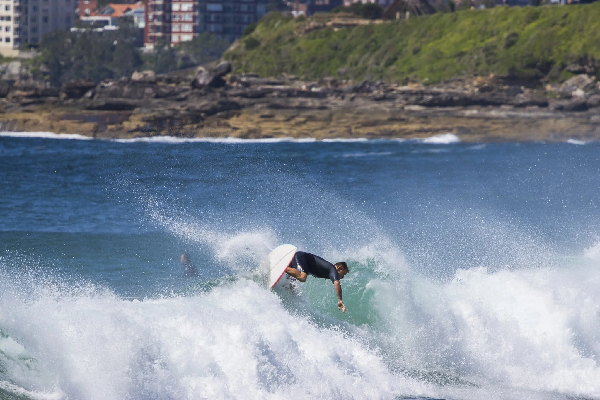 A person surfing on a wave near the shoreline with buildings and greenery in the background.
