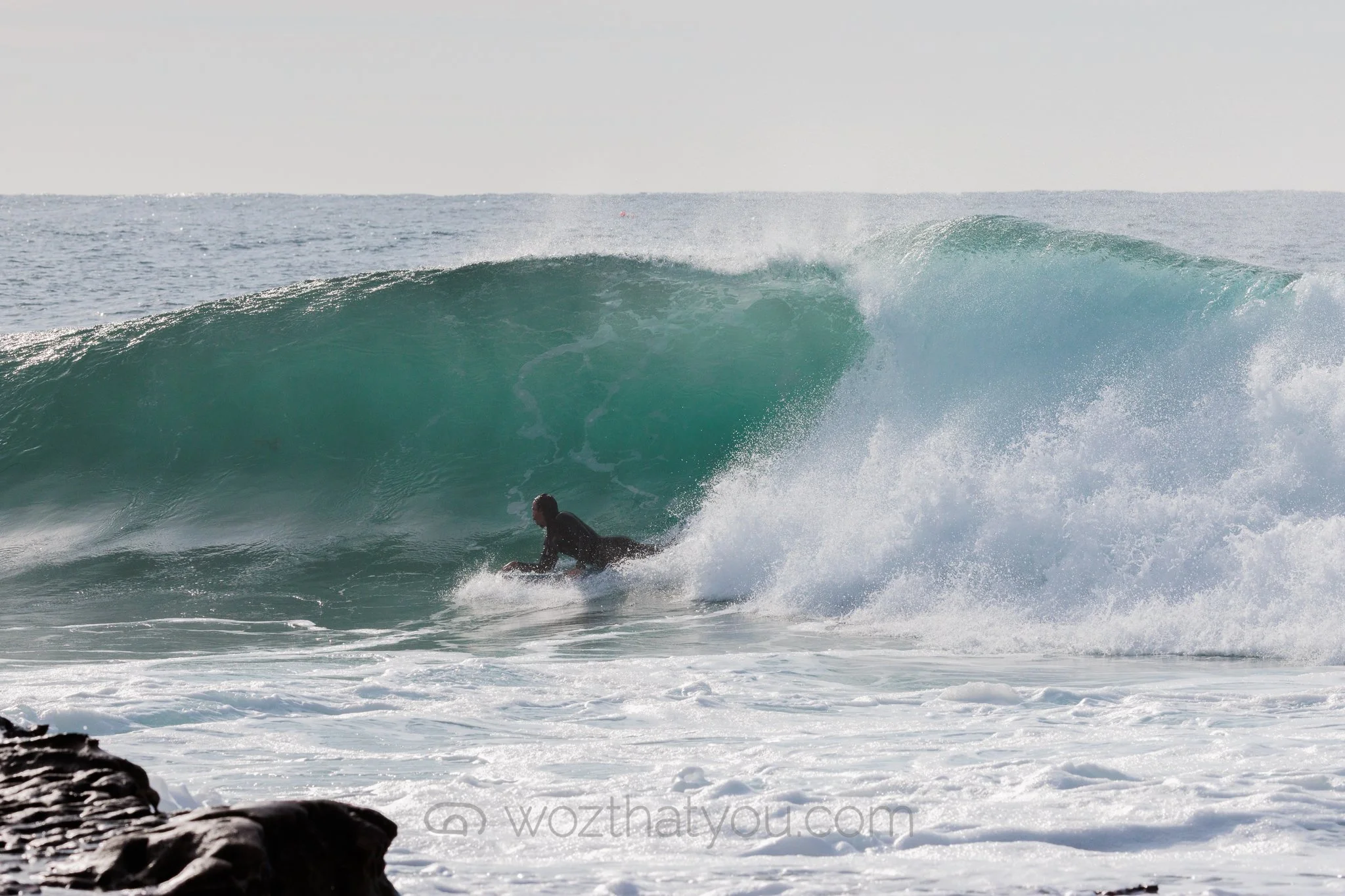 A surfer in a black wetsuit riding inside the barrel of a large ocean wave with foam and spray, with a rocky shoreline in the foreground.