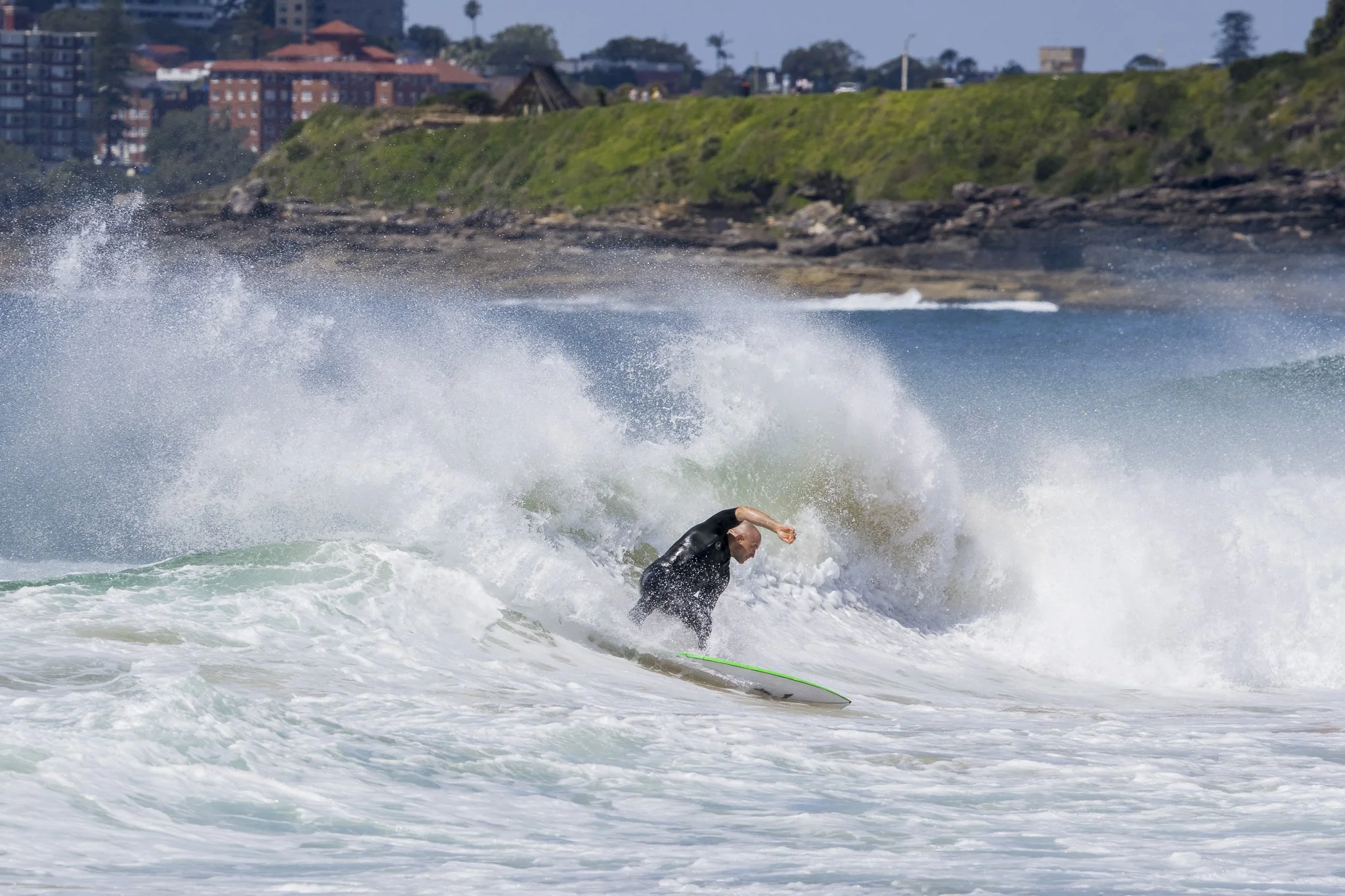 A person surfing on a wave near the shoreline, with green hills and buildings in the background.
