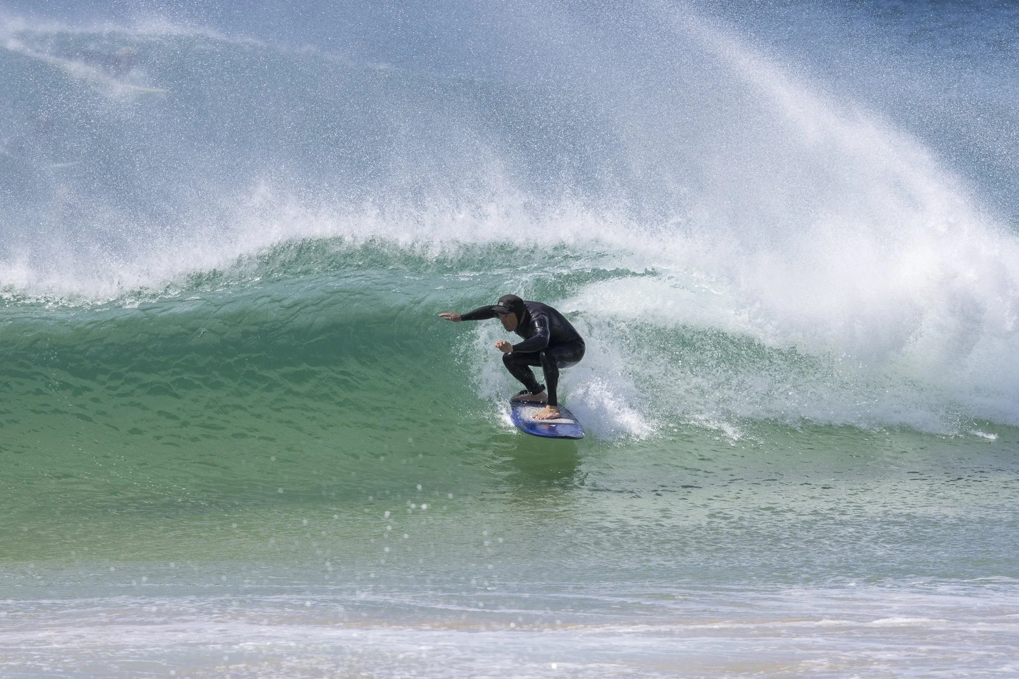 A person dressed in a black wetsuit and helmet surfing on a blue surfboard on a green wave at the beach.