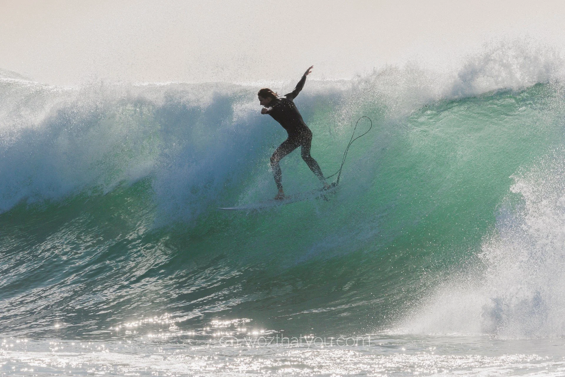 A person is surfing on a wave in the ocean, wearing a black wetsuit and holding a surfboard leash.
