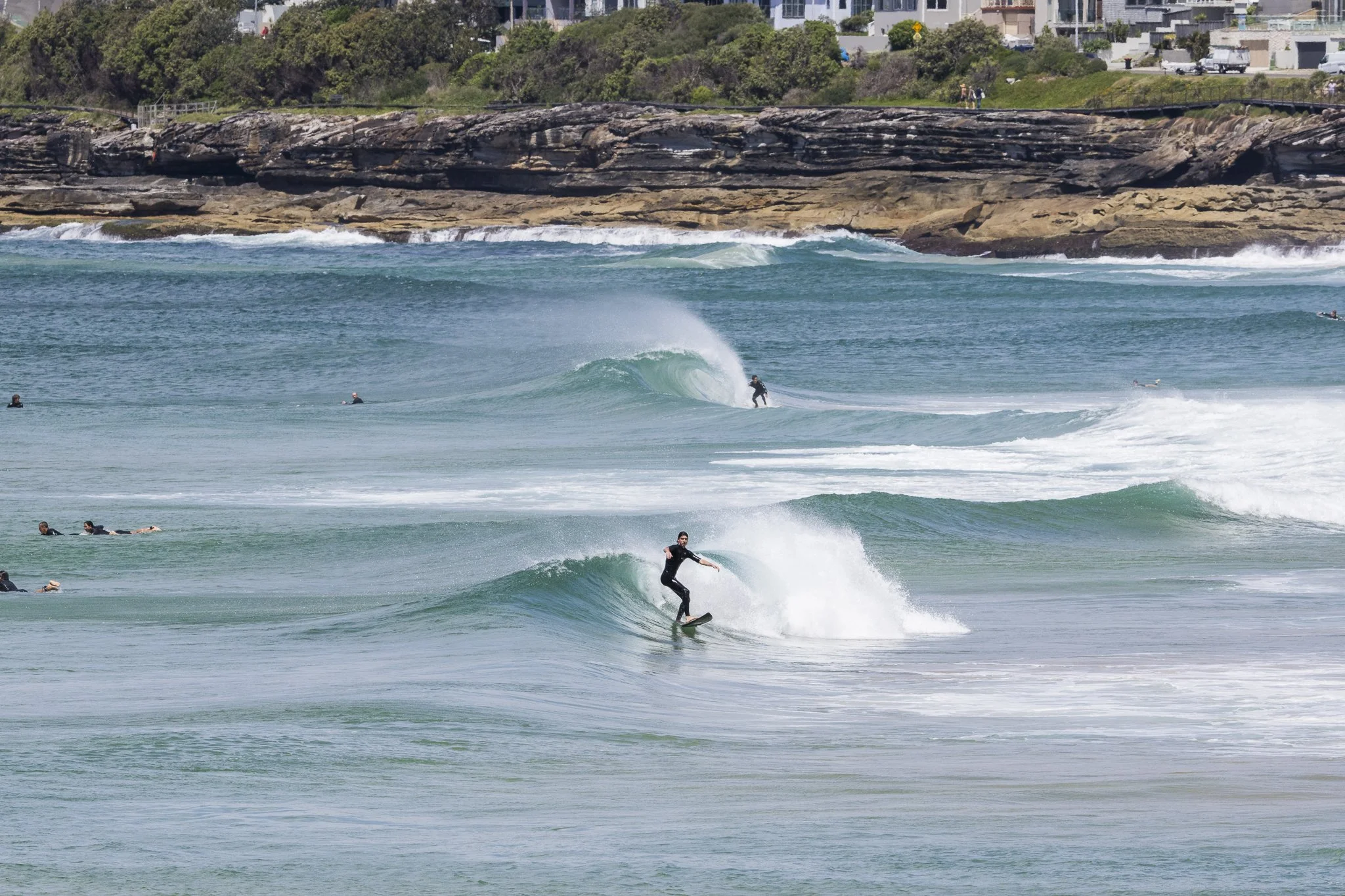 People surfing and swimming in the ocean near a rocky shoreline with houses in the background.
