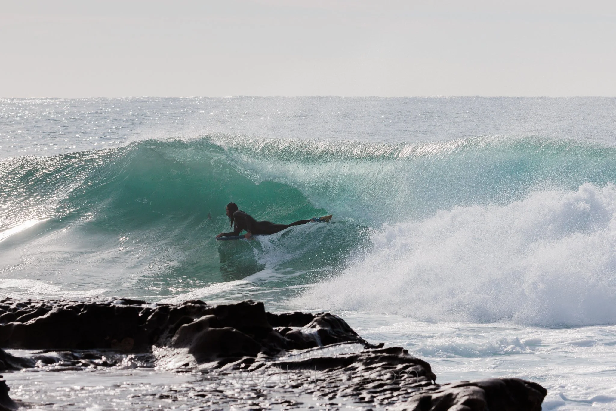A person in a wetsuit riding a surfboard on a wave near the rocky shoreline.