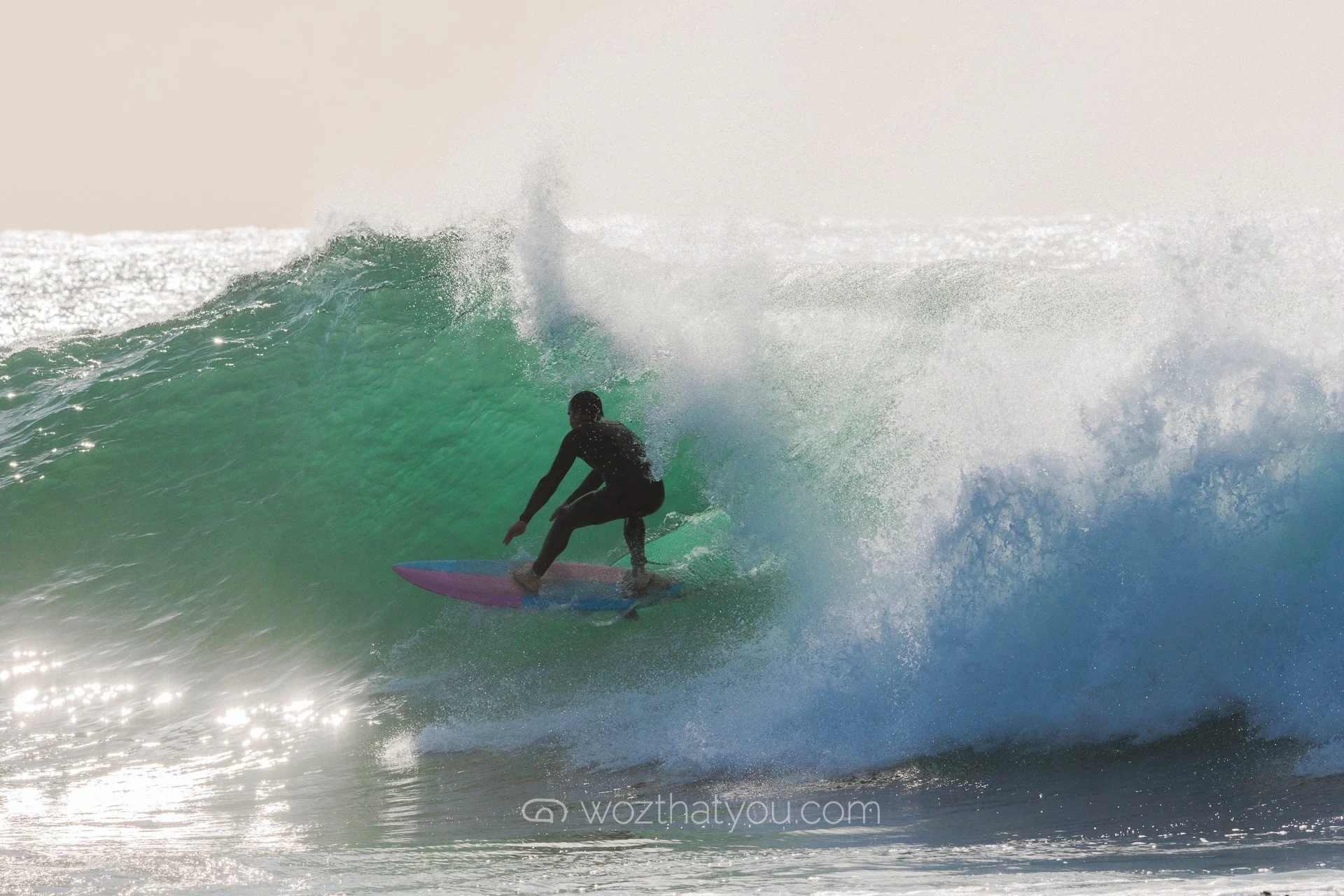 Surfer rides a wave in the ocean, creating a splash with sunlight reflecting off the water.
