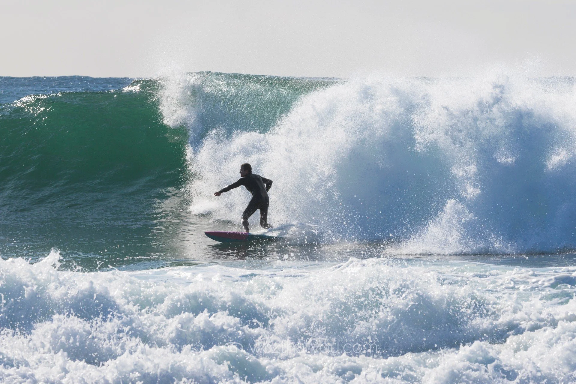 A person surfing on a large ocean wave with white foam and spray behind them.