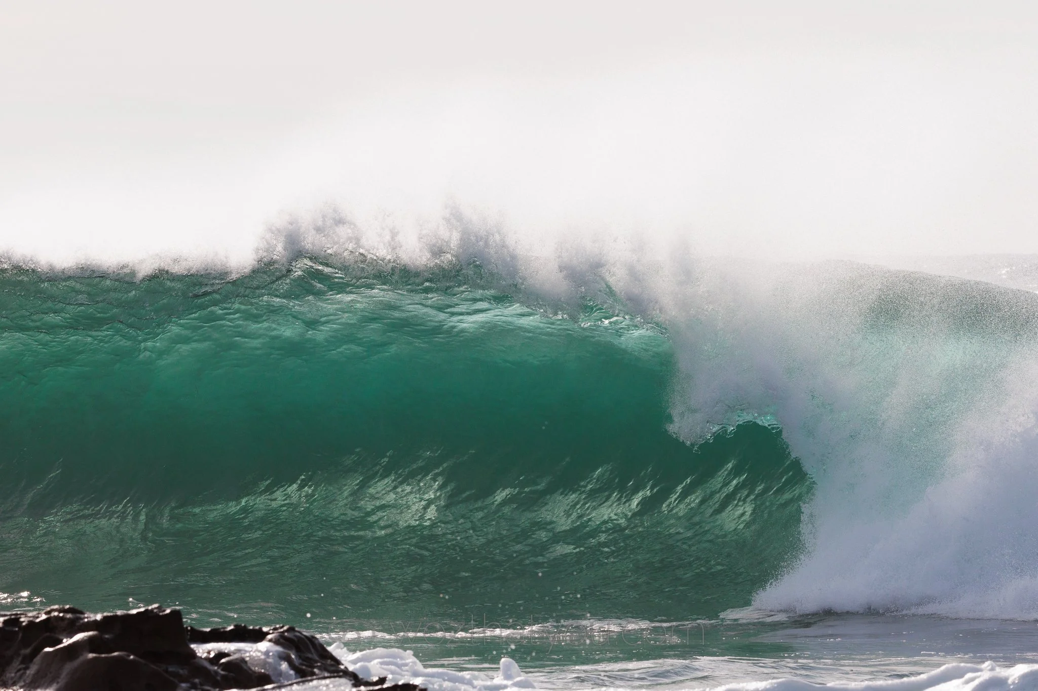 Large green ocean wave crashing near rocks in the foreground, white spray and mist in the air.