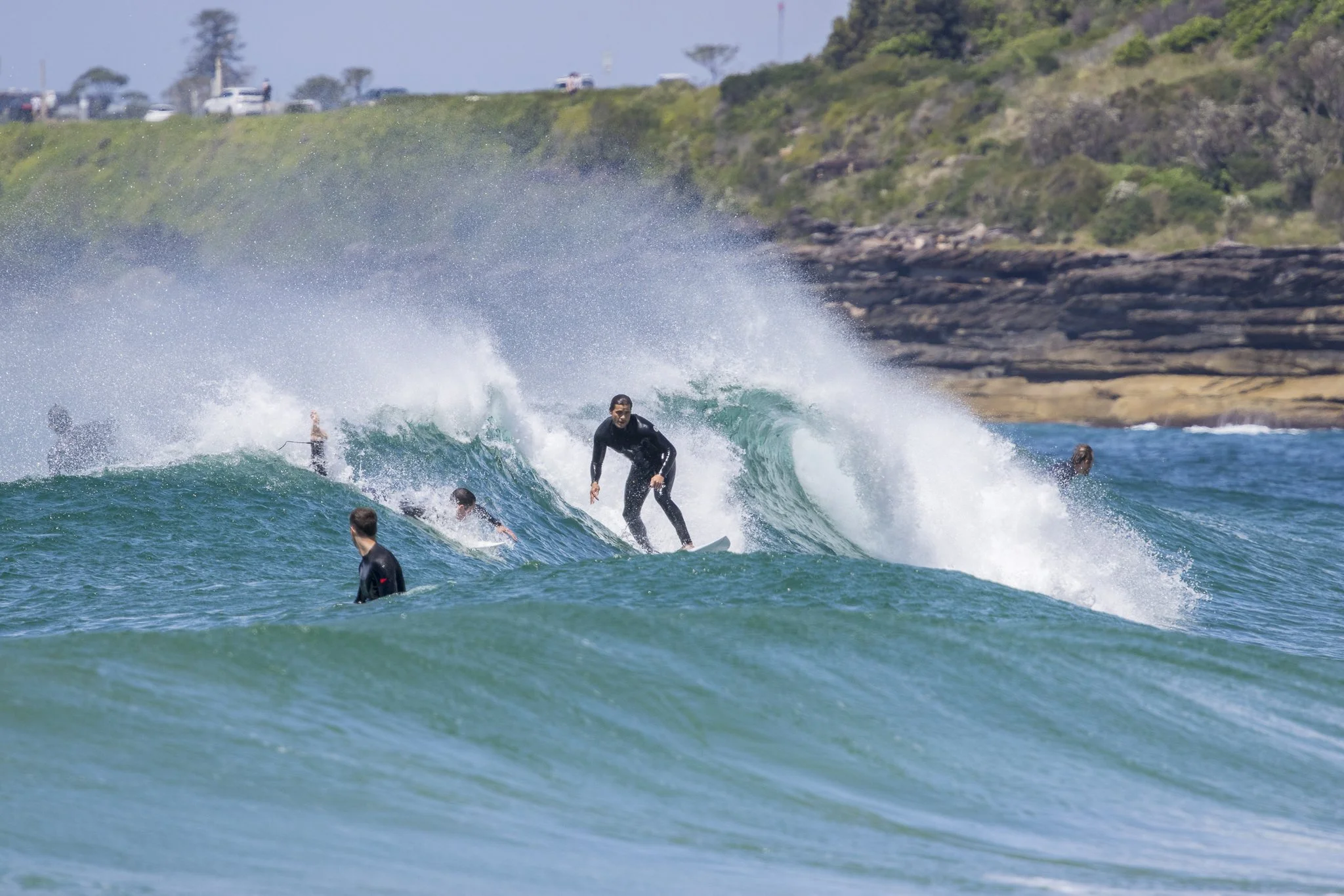 Surfers riding and waiting for waves at the beach with rocky cliffs and cars on a road in the background.