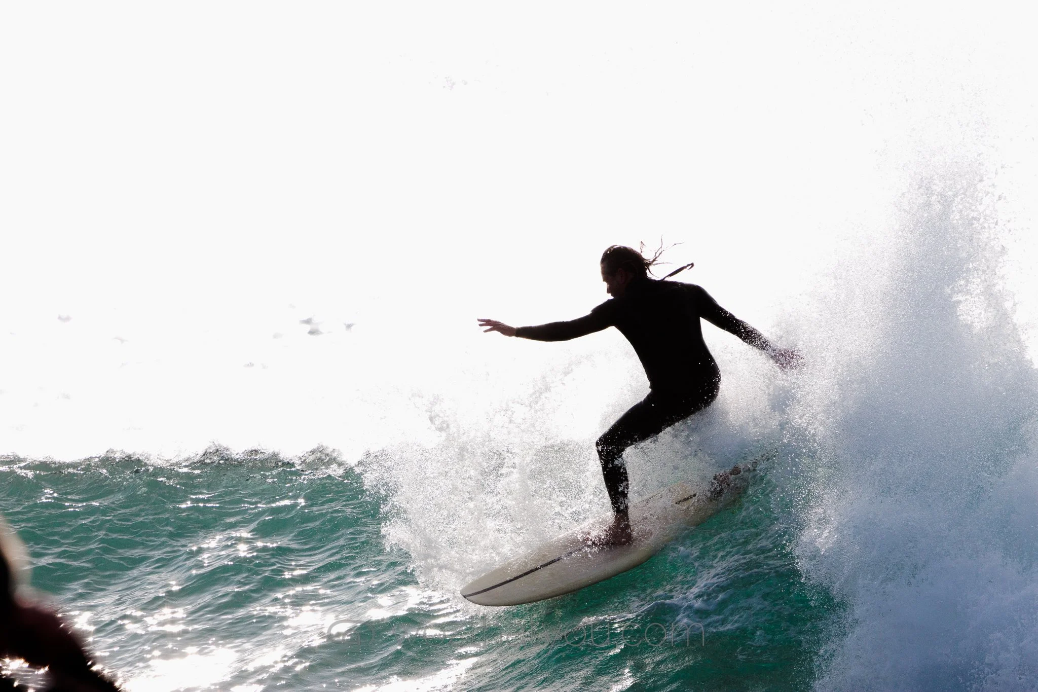 A silhouette of a person surfing on a wave with spray of water around them on a sunny day.