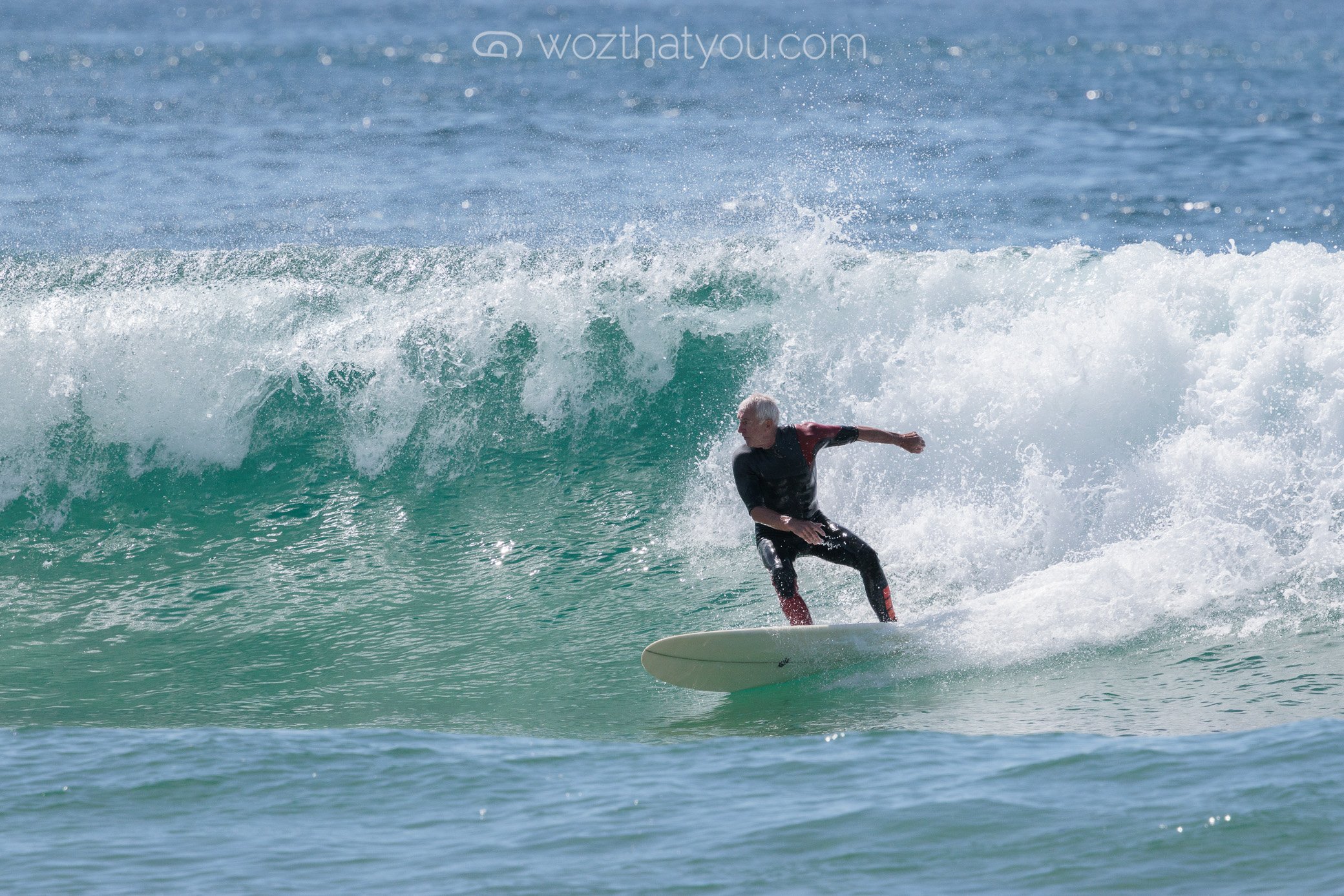 A person with gray hair wearing a black wetsuit surfing on a white surfboard on a wave in the ocean.