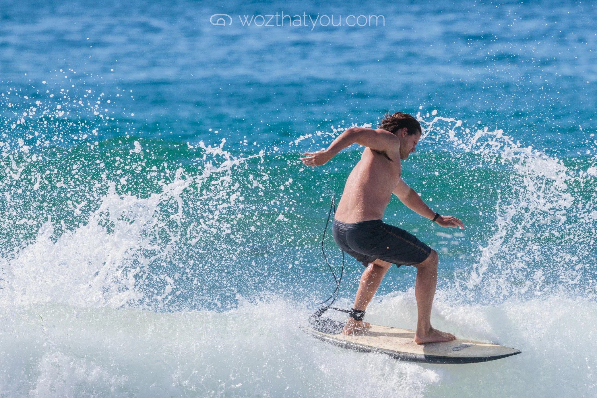A man with long hair surfing on a wave in the ocean wearing black shorts, water splashing around him.