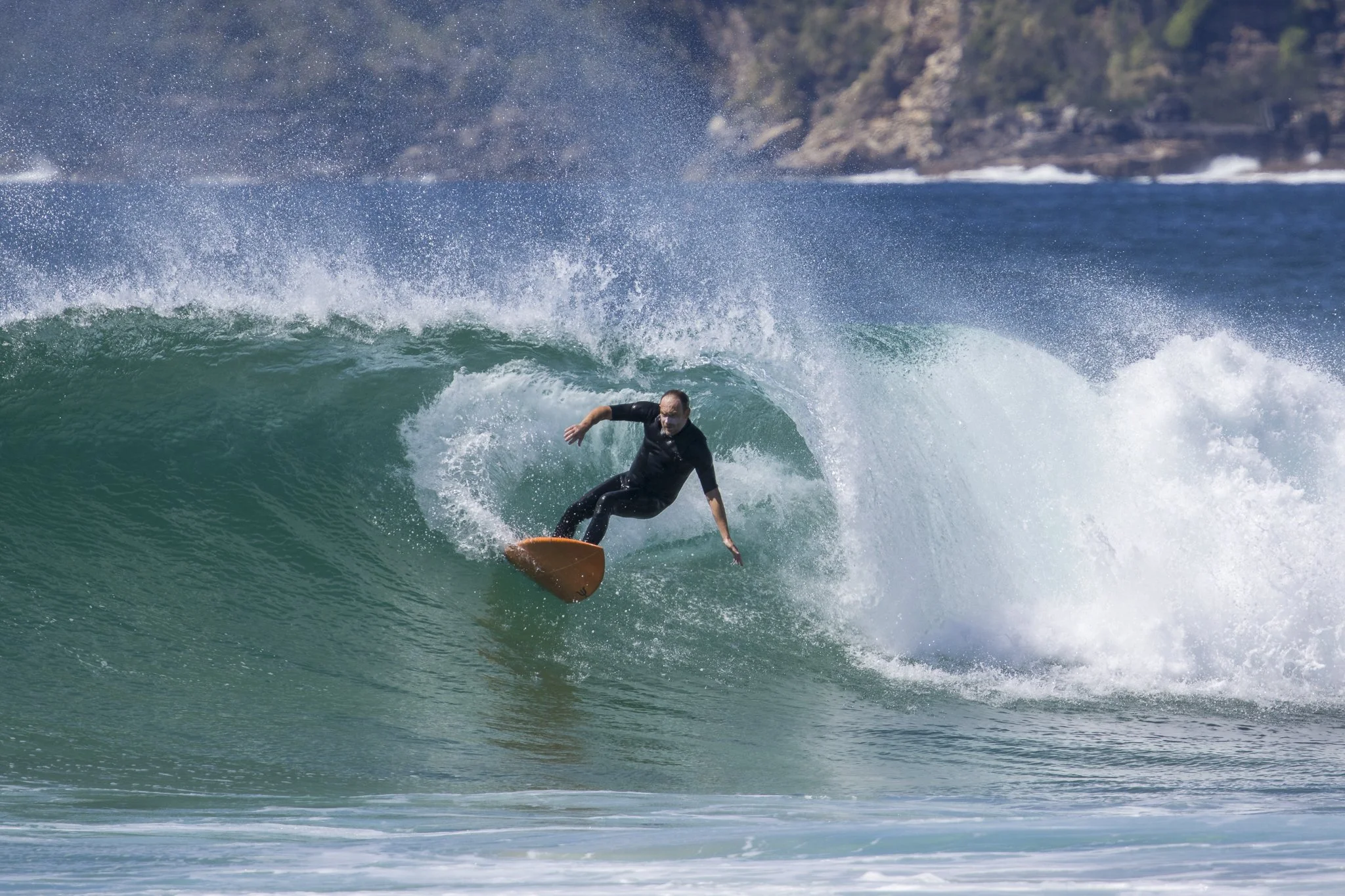 A man surfing on a large green wave with a rocky shoreline in the background.