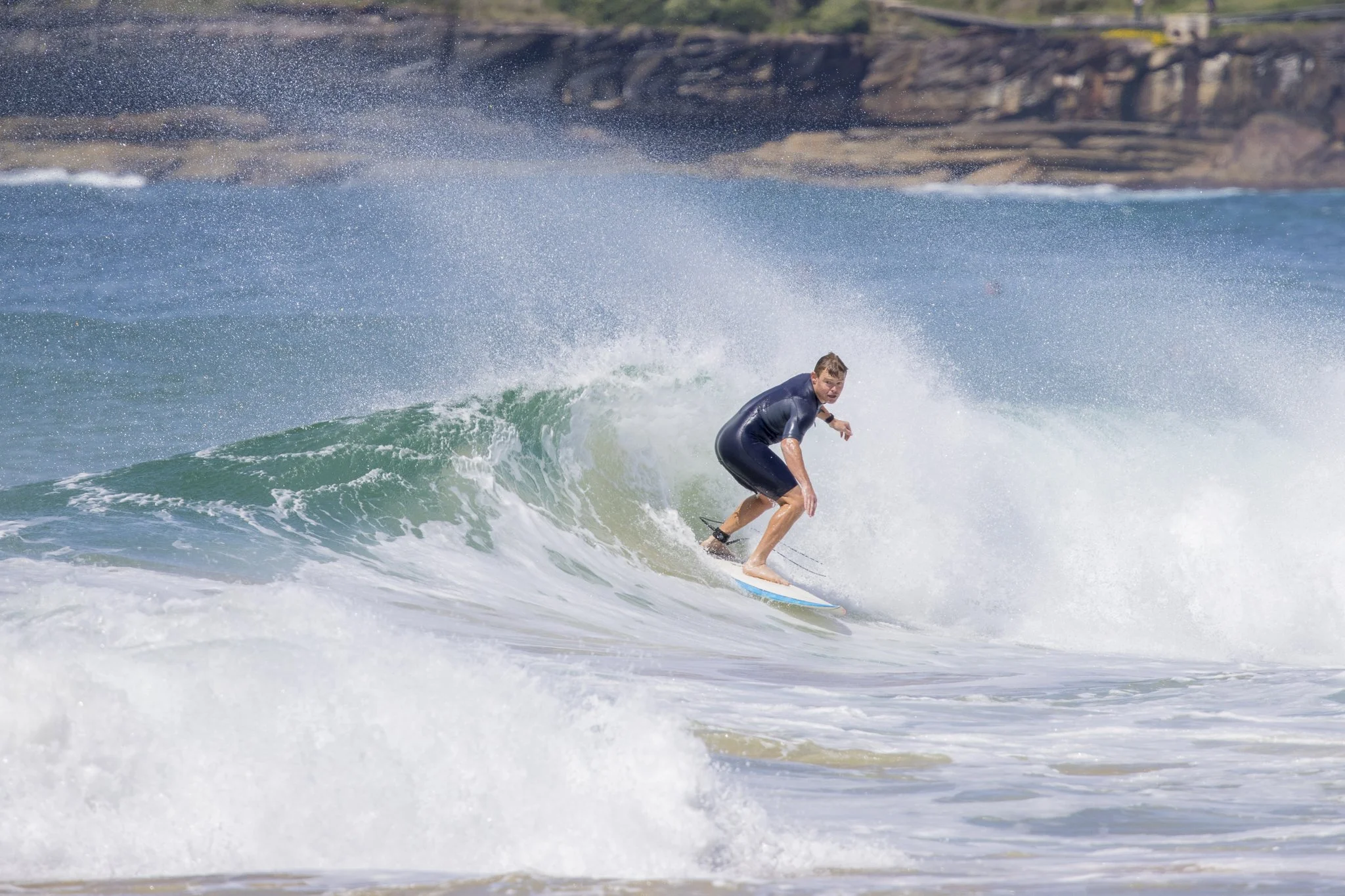 A young man in a black wetsuit surfing on a wave at the beach with rocky cliffs in the background.