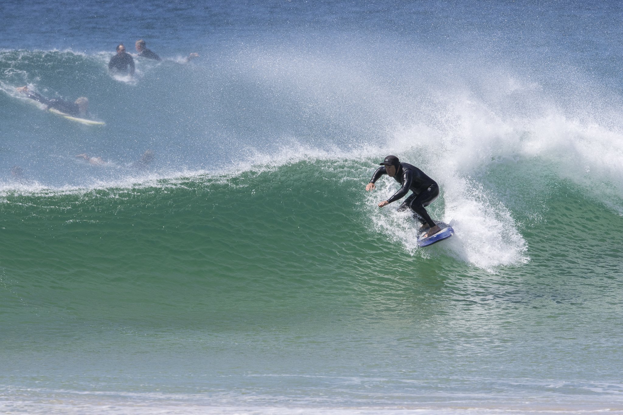 A person in a black wetsuit surfing on a large green wave, with several other surfers in the background.