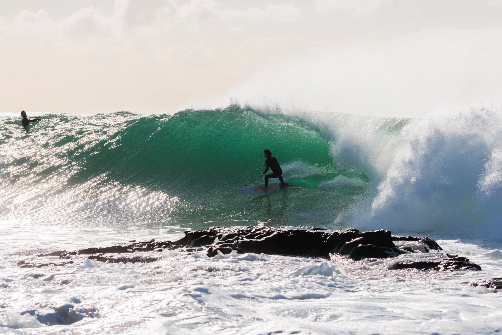A person surfing on a large green wave near rocky shoreline, with another surfer visible in the distance.