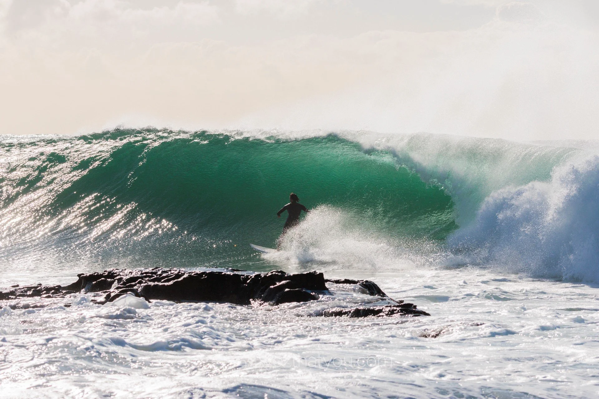 A person surfing on a large green wave with white foam in the foreground and pastel sky in the background.