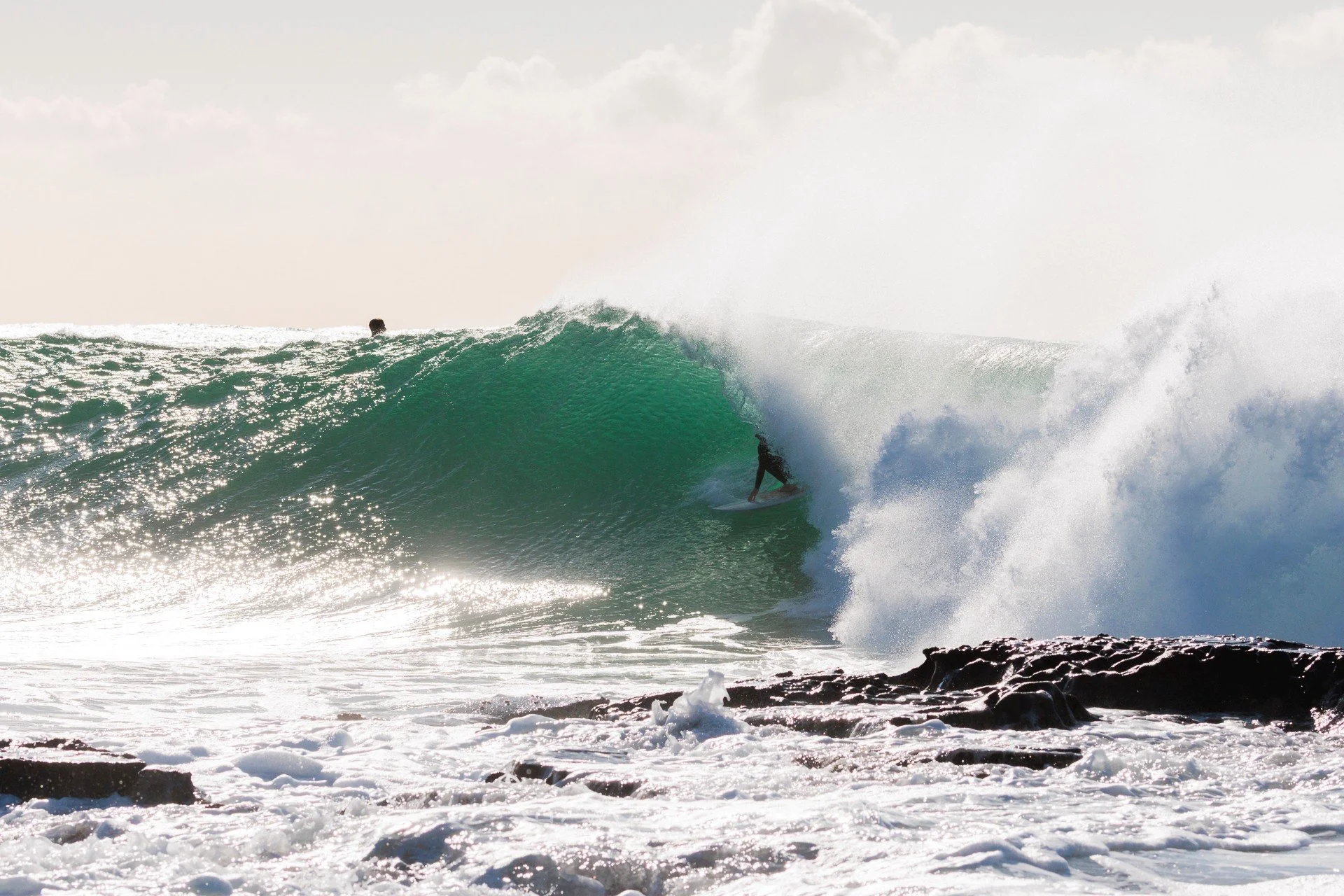 A person surfing a large wave near a rocky shoreline with another person in the distance in the water.