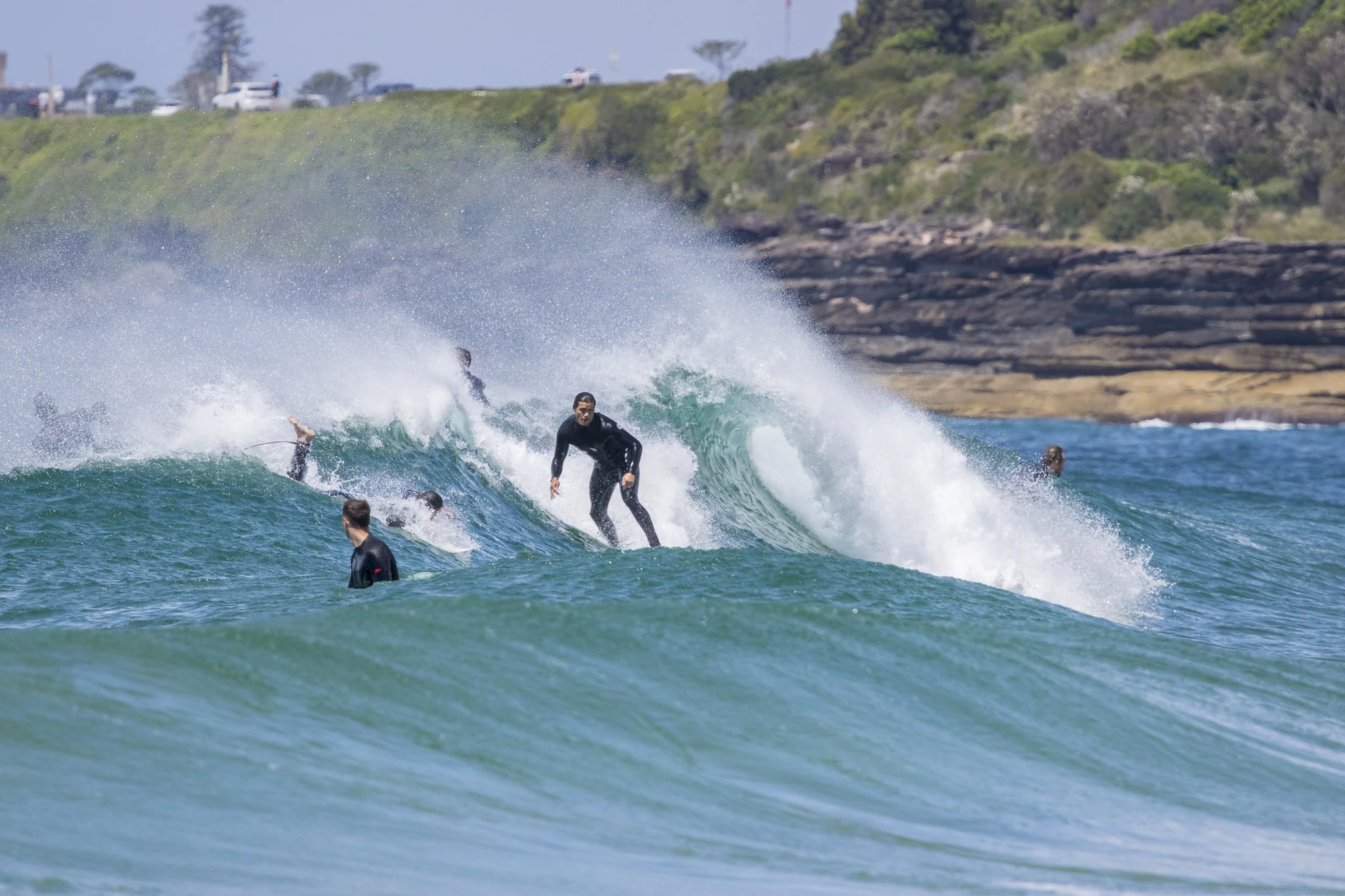 Group of people surfing on ocean waves near rocky shoreline, with some in the water and one standing on a surfboard riding a wave.