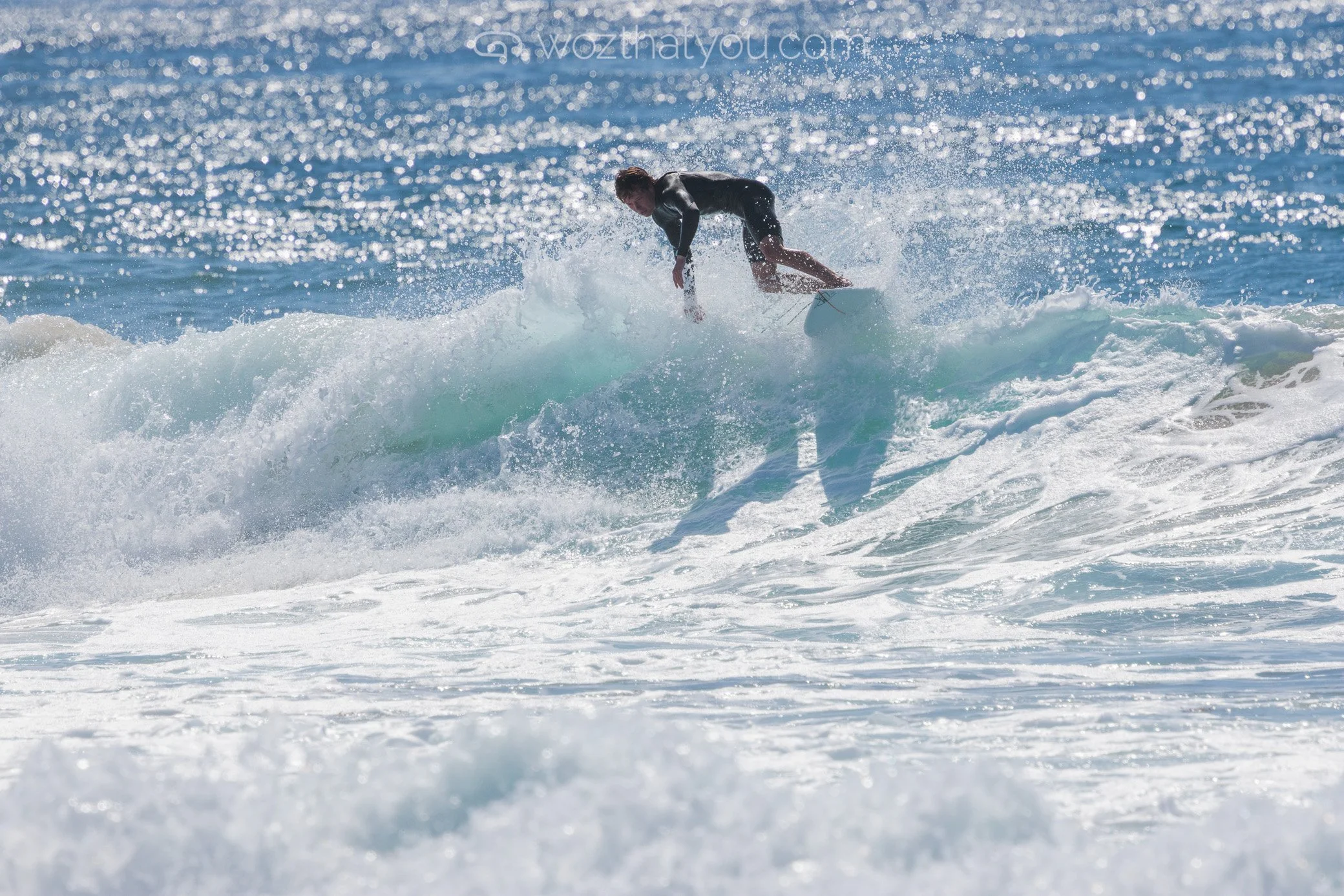 A person surfing on a wave in the ocean, wearing black wetsuit, with water splashing around.