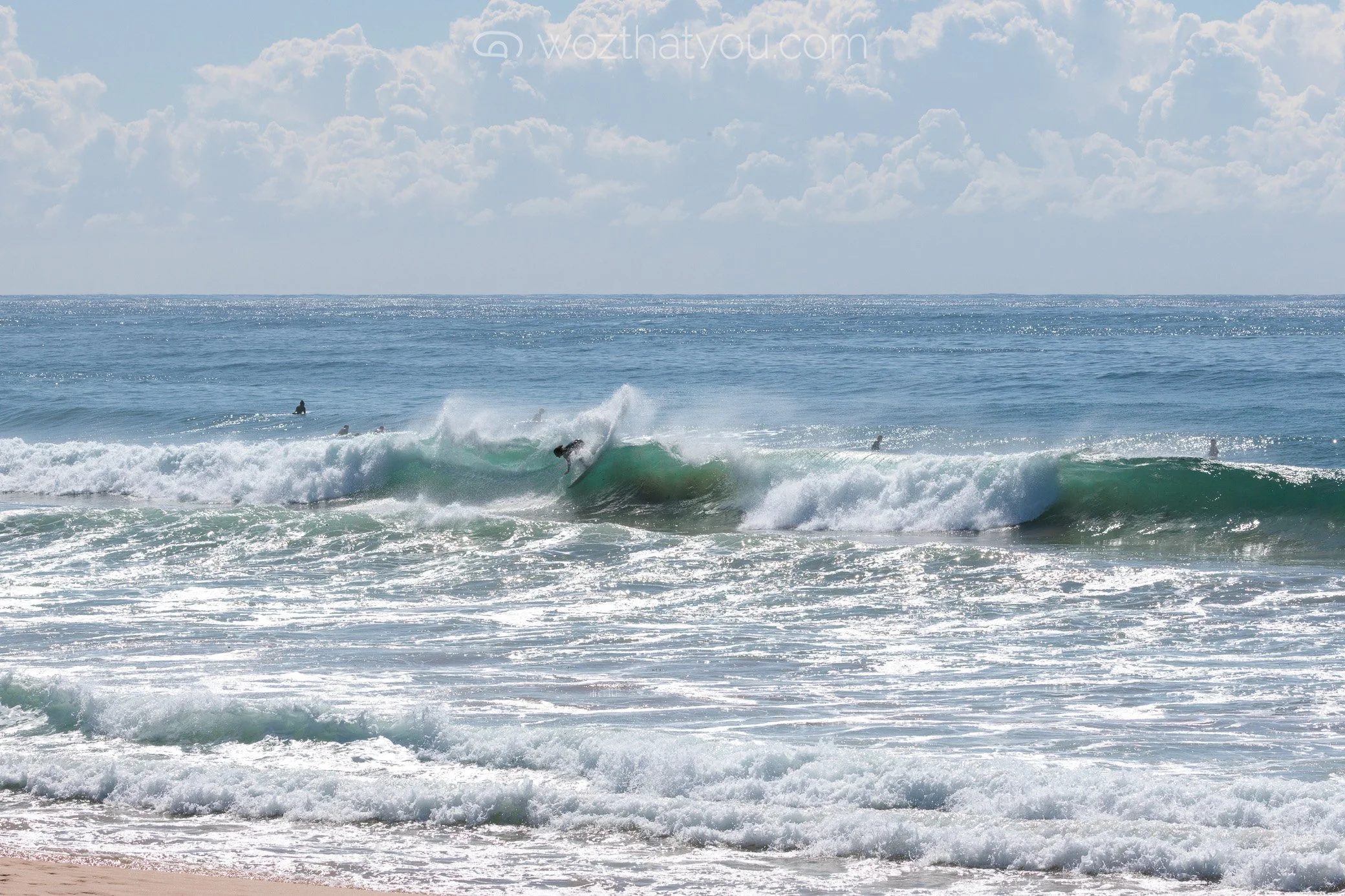 Surfer riding a wave at the beach on a sunny day with a partly cloudy sky, and other surfers in the water.