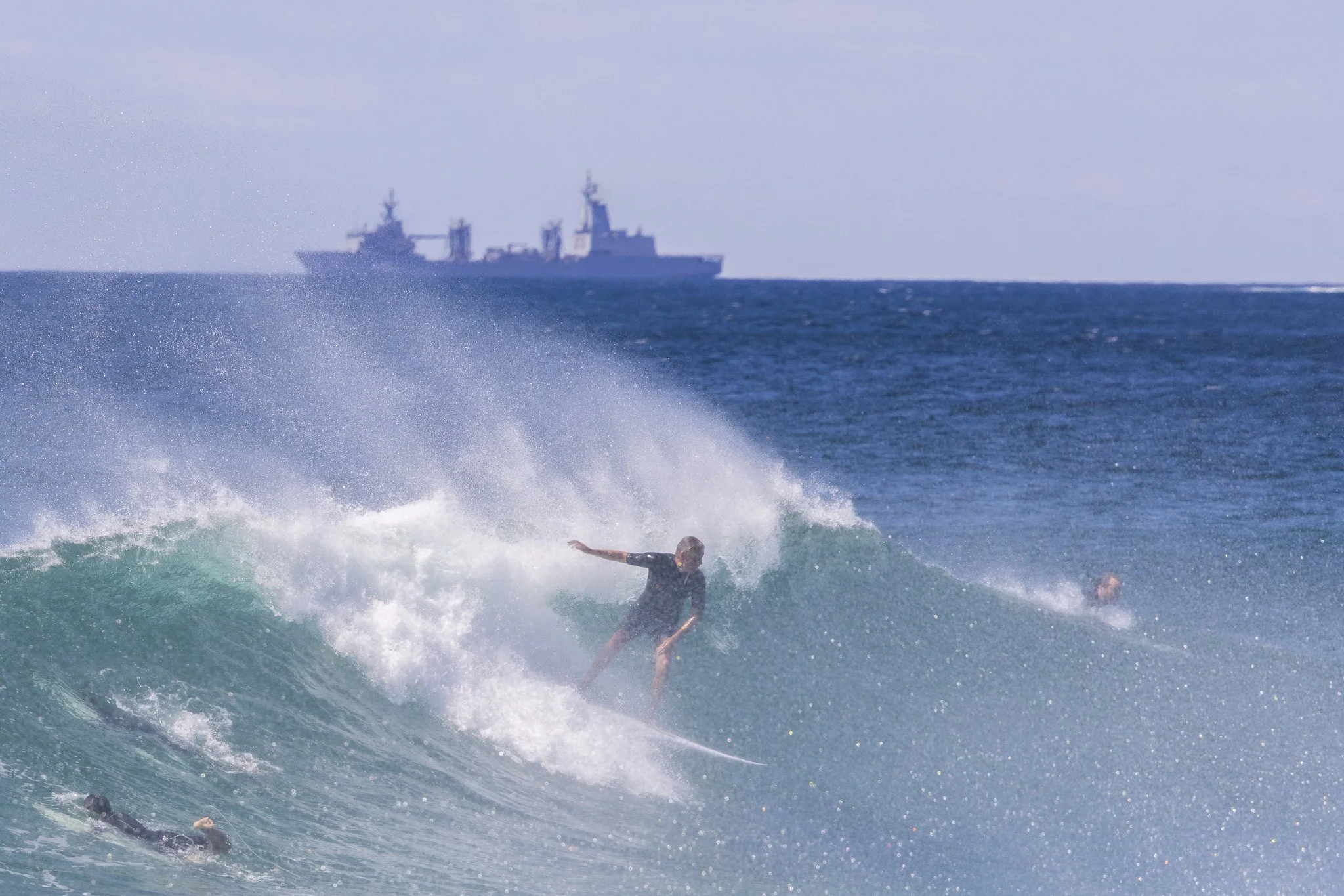 A person surfing on a wave in the ocean with a military ship on the horizon in the background.