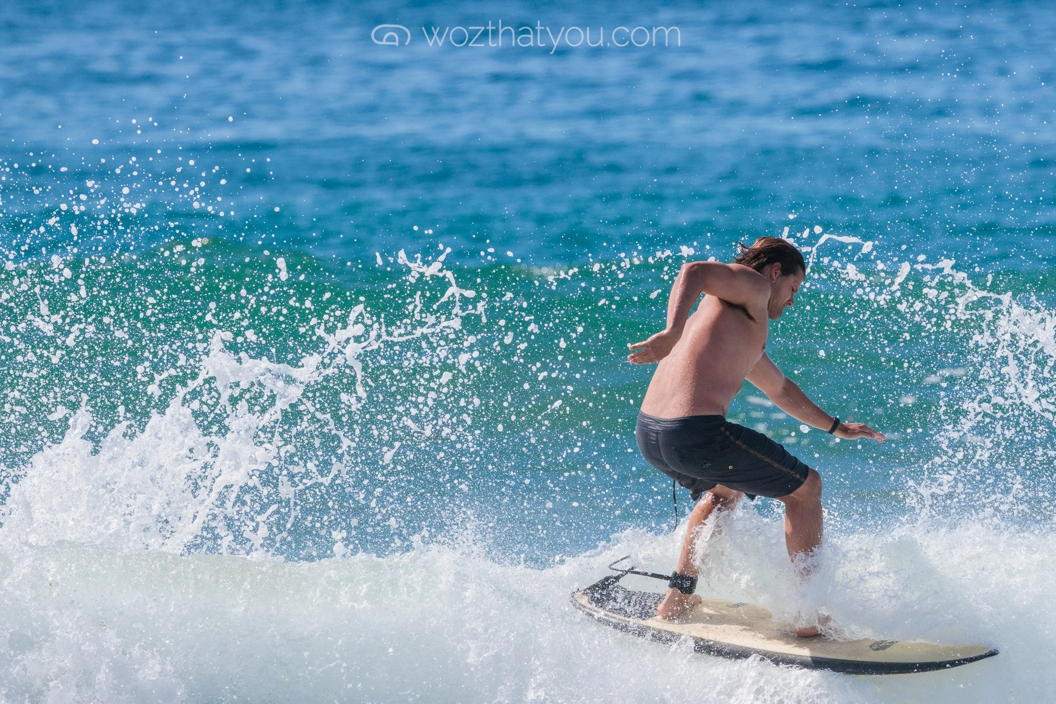 A man with long hair is surfing on a wave in the ocean, wearing black shorts and no shirt.