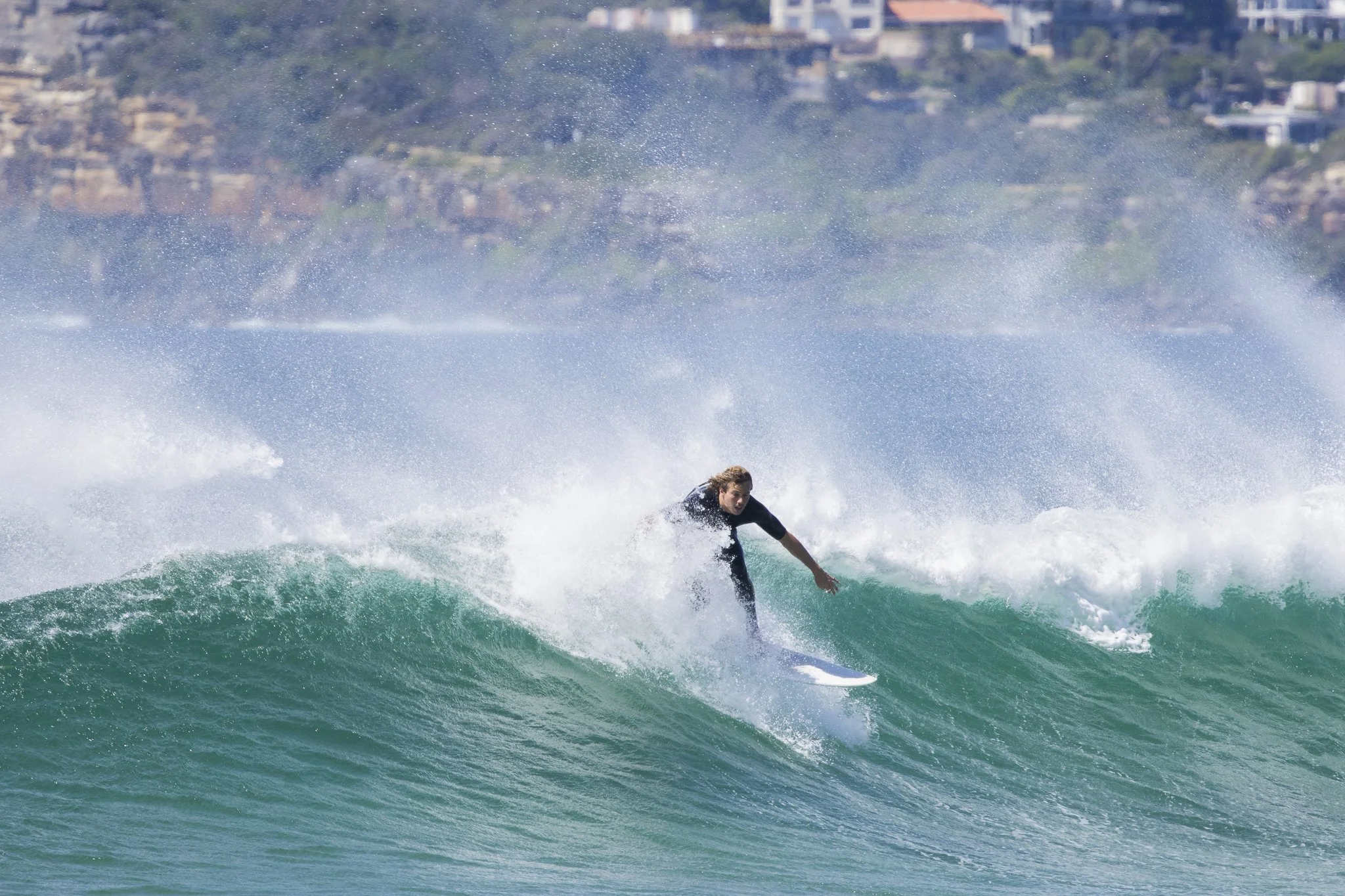A person surfing on a wave near the coast with a cityscape in the background.