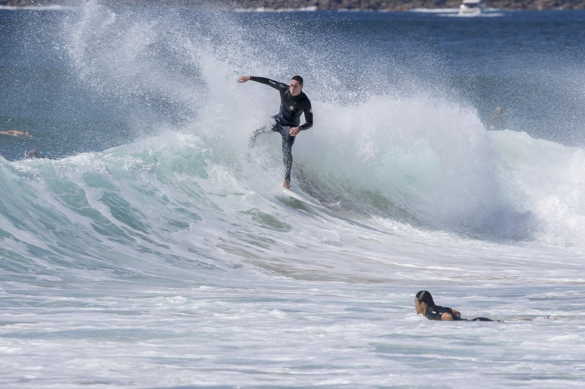 A male and female surfer riding a wave in the ocean, with the male standing on his surfboard and the female paddling in the water nearby.