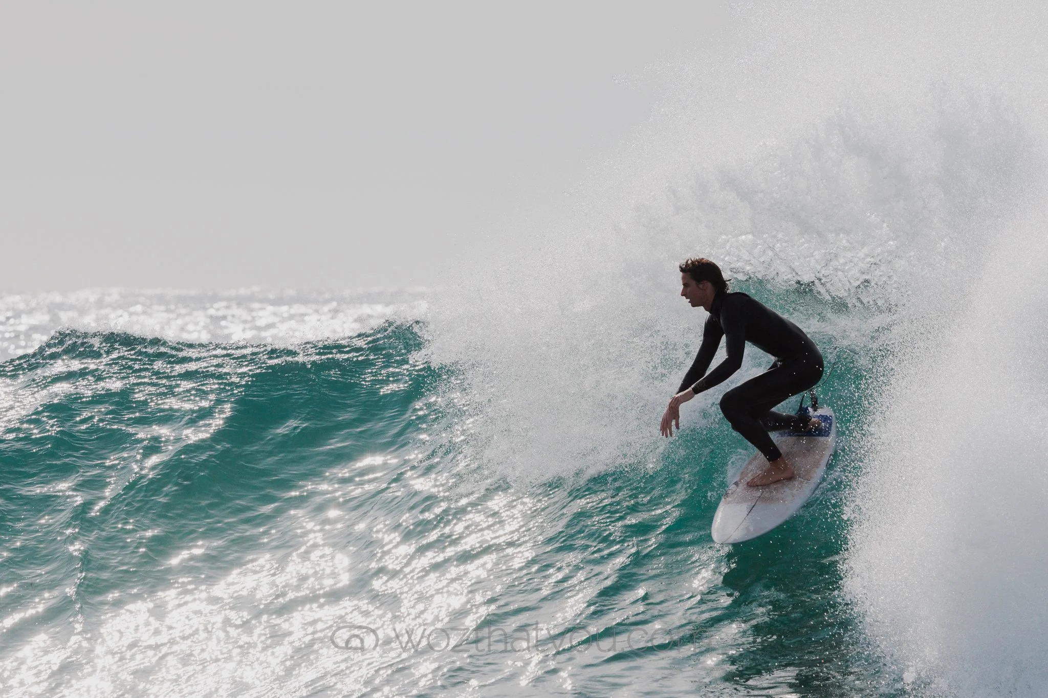 A person in a black wetsuit surfing on a wave with sunlit water and spray in the air.
