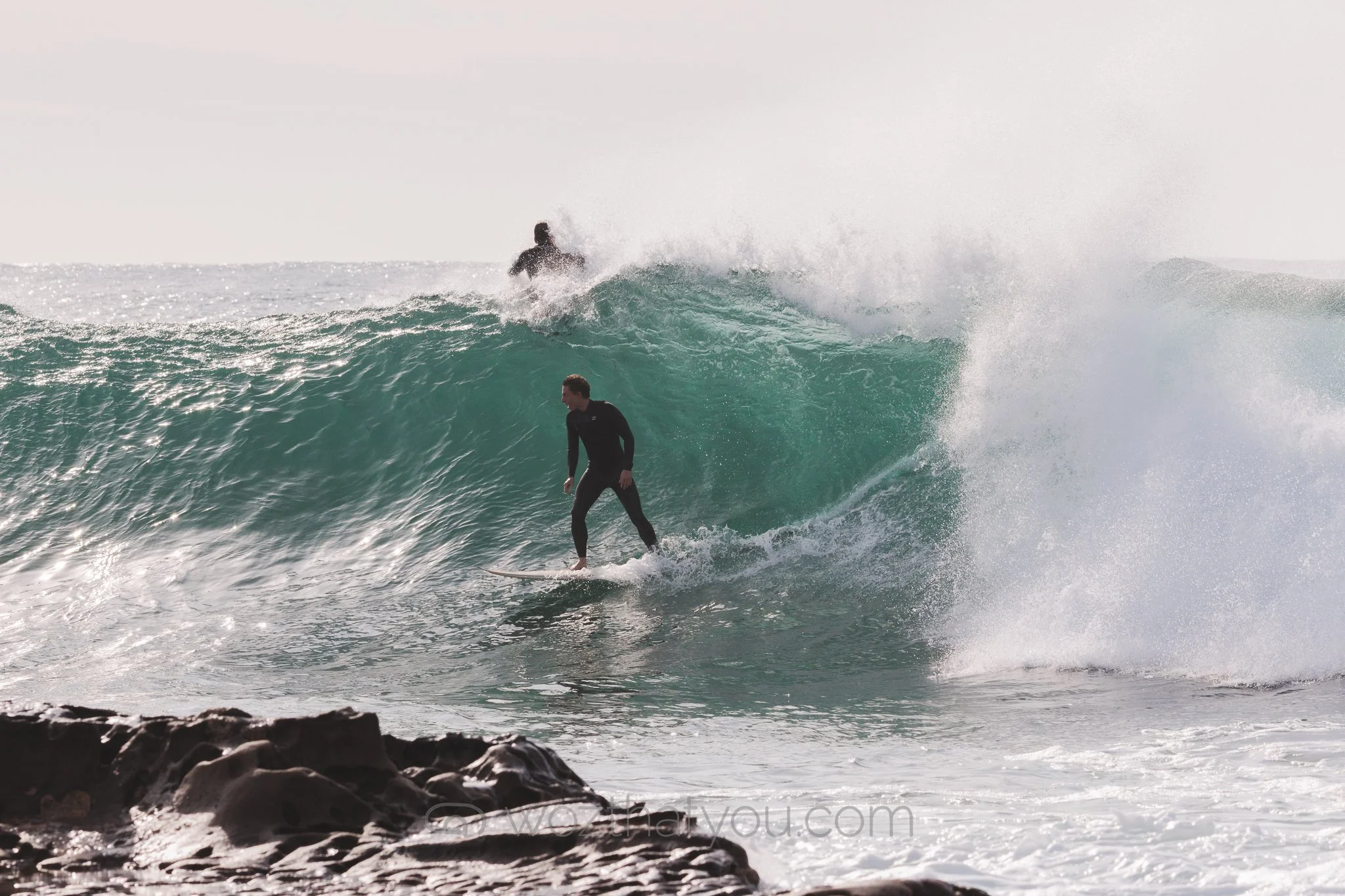 A person surfing on a wave with another surfer riding in the background.