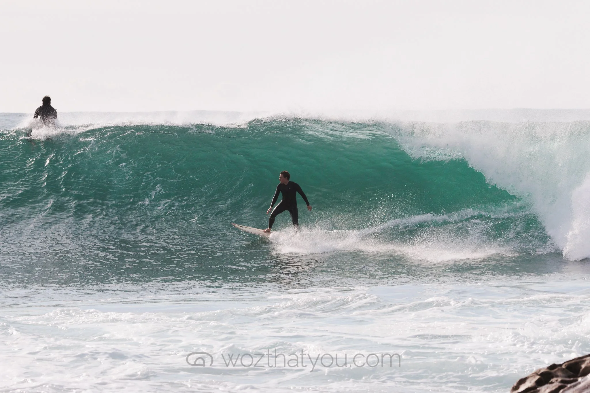 Surfer riding a wave with another surfer in the background near the ocean shoreline.
