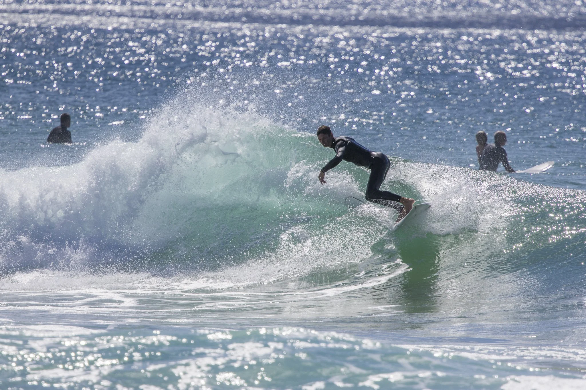 A person surfing on a wave in the ocean while wearing a black wetsuit, with two other surfers visible in the background.