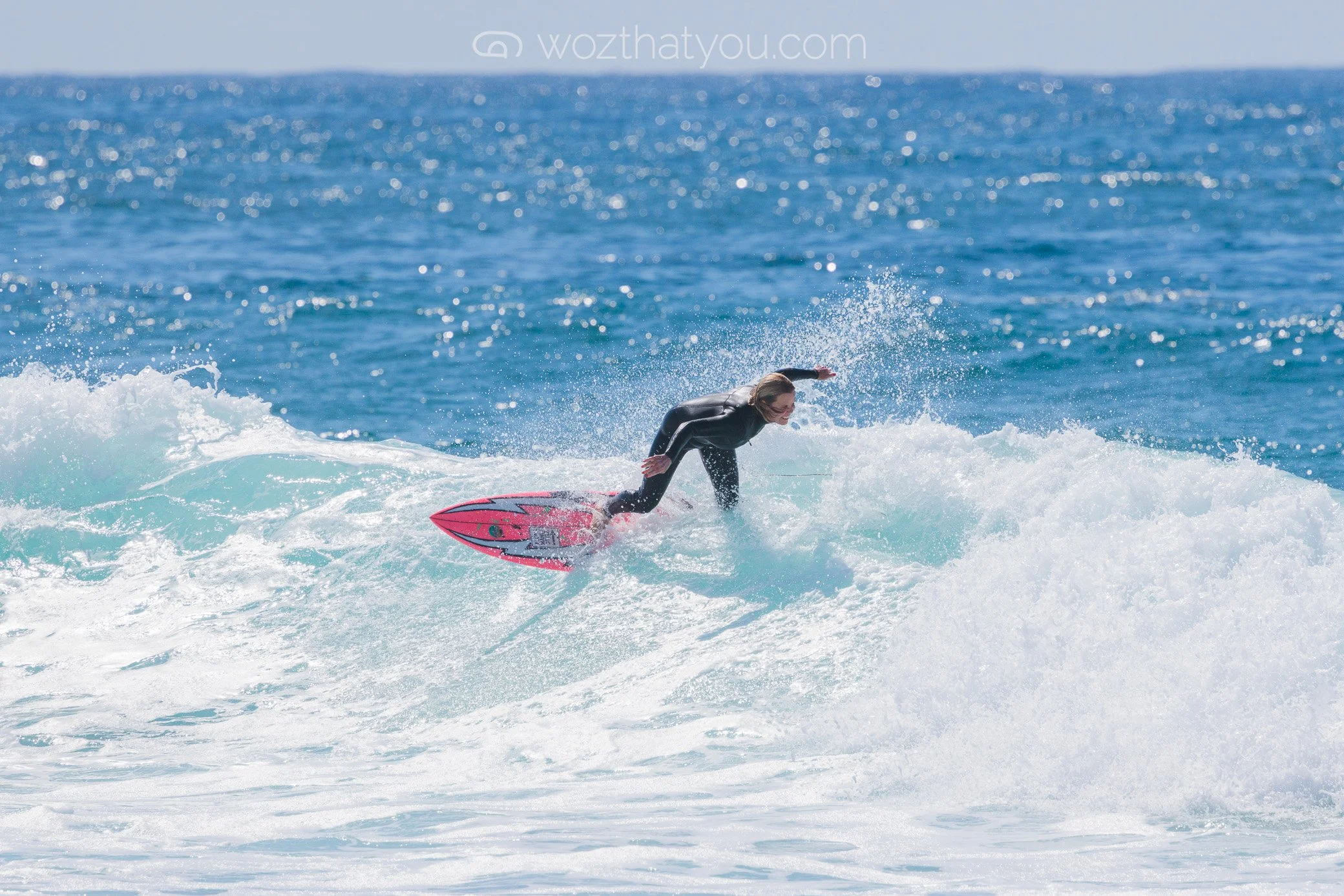 A woman in a black wetsuit surfing on a pink surfboard in the ocean on a sunny day.