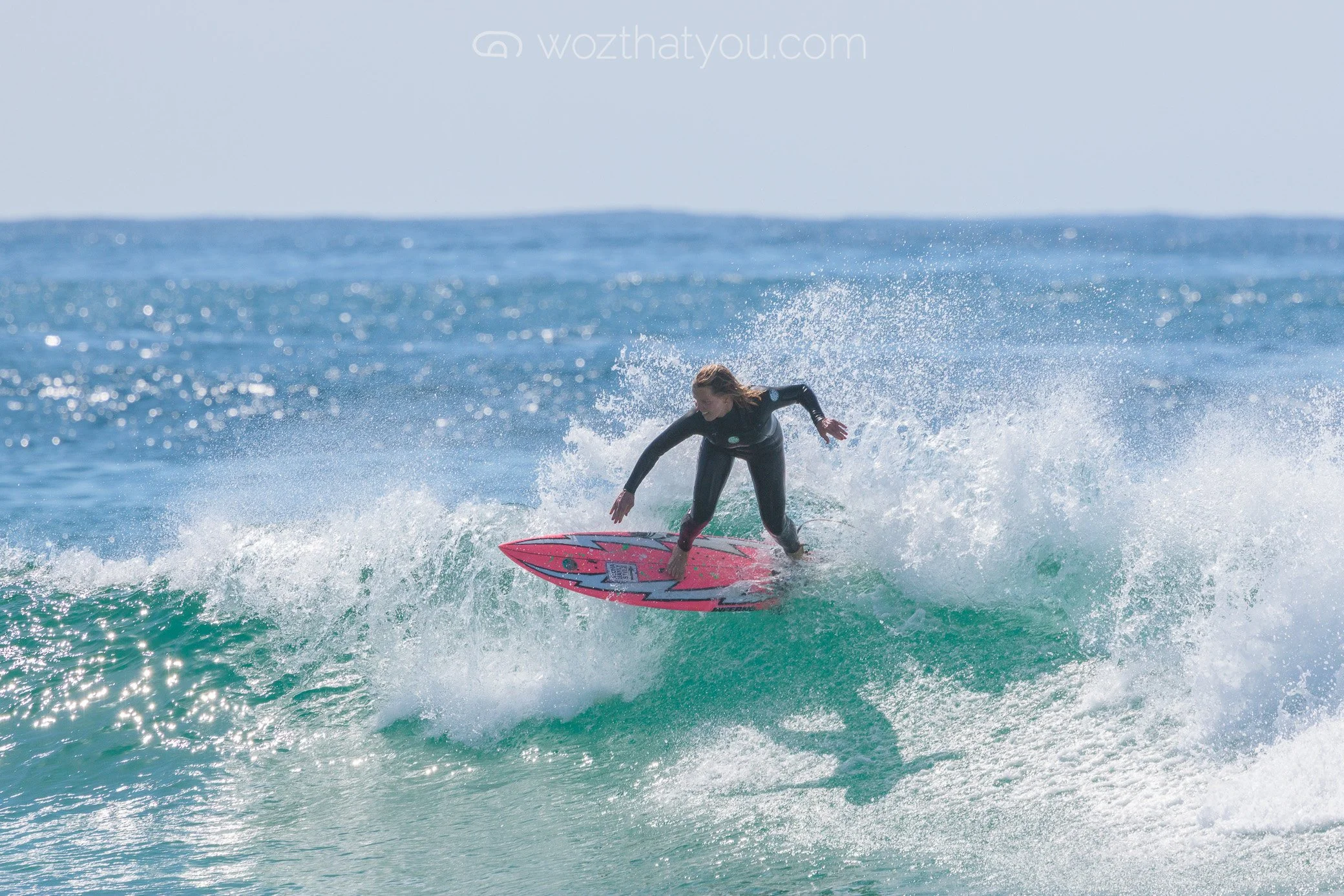 A woman surfing on a pink surfboard in the ocean, wearing a black wetsuit, with waves and water spray around her.