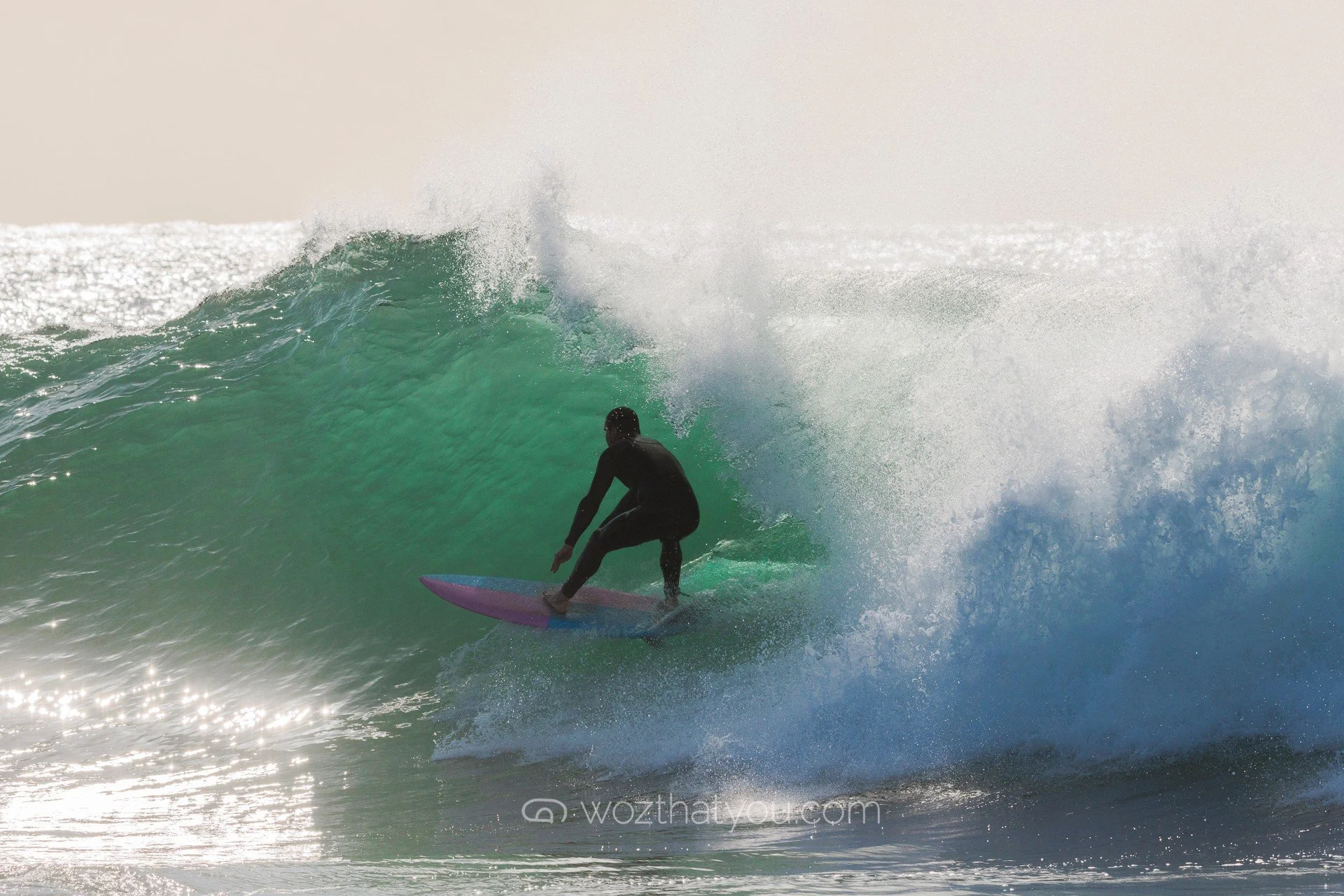 A person in a black wetsuit surfing on a pink and blue surfboard on a large green wave with white spray, during daytime.