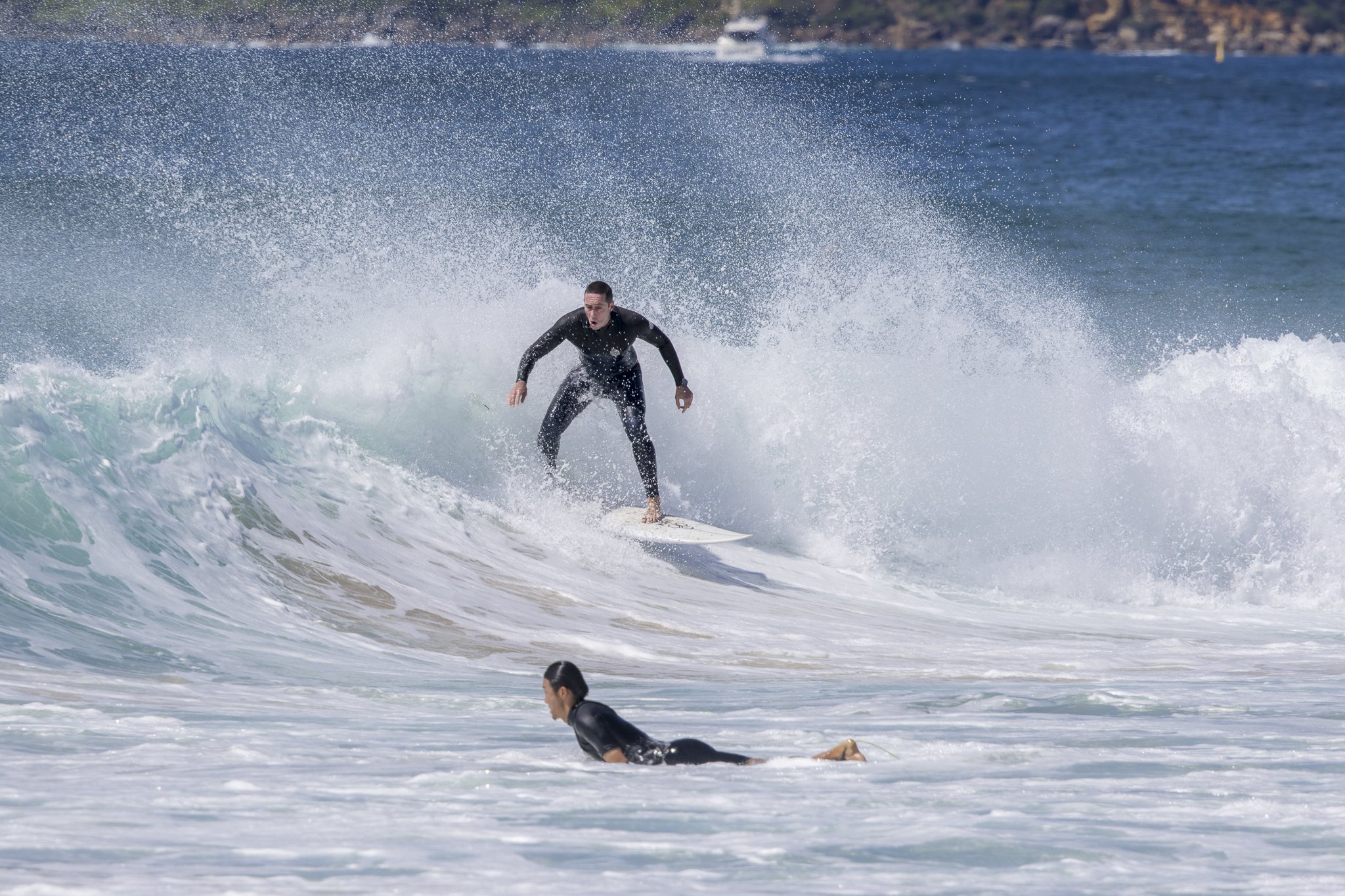 A man in a wetsuit surfing on a wave while another person lies on the water nearby.
