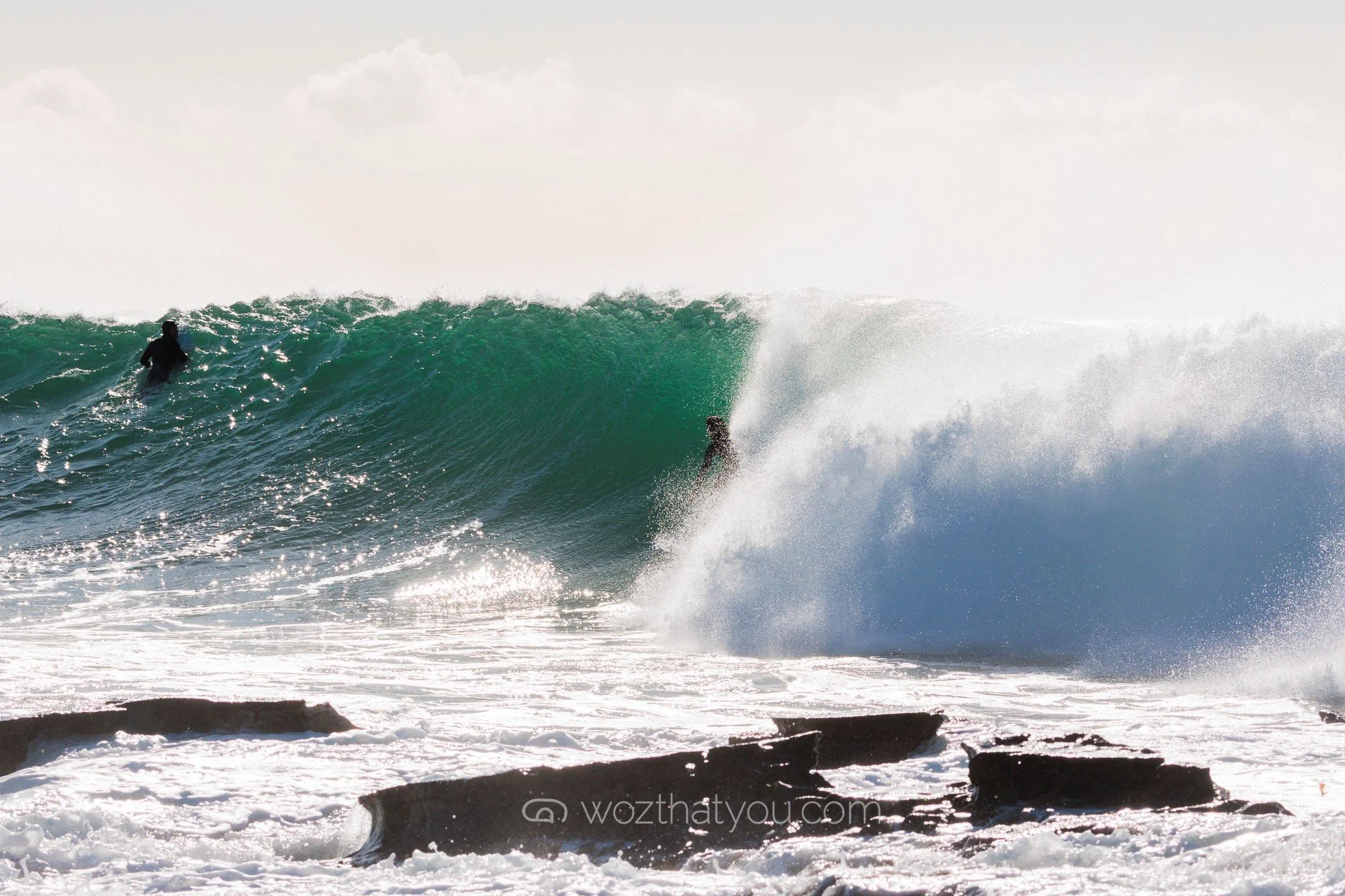 Two surfers riding a large ocean wave near the shoreline with rocks in the foreground.