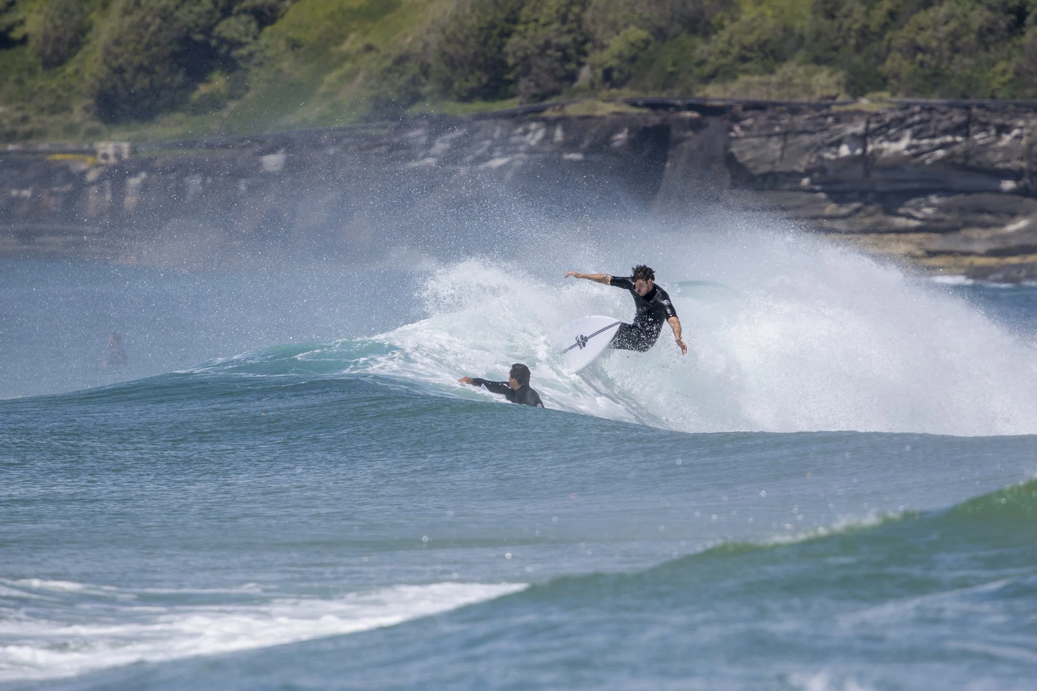 A surfer executing a turn on a wave with another person lying on a surfboard nearby, with a rugged shoreline and green hills in the background.