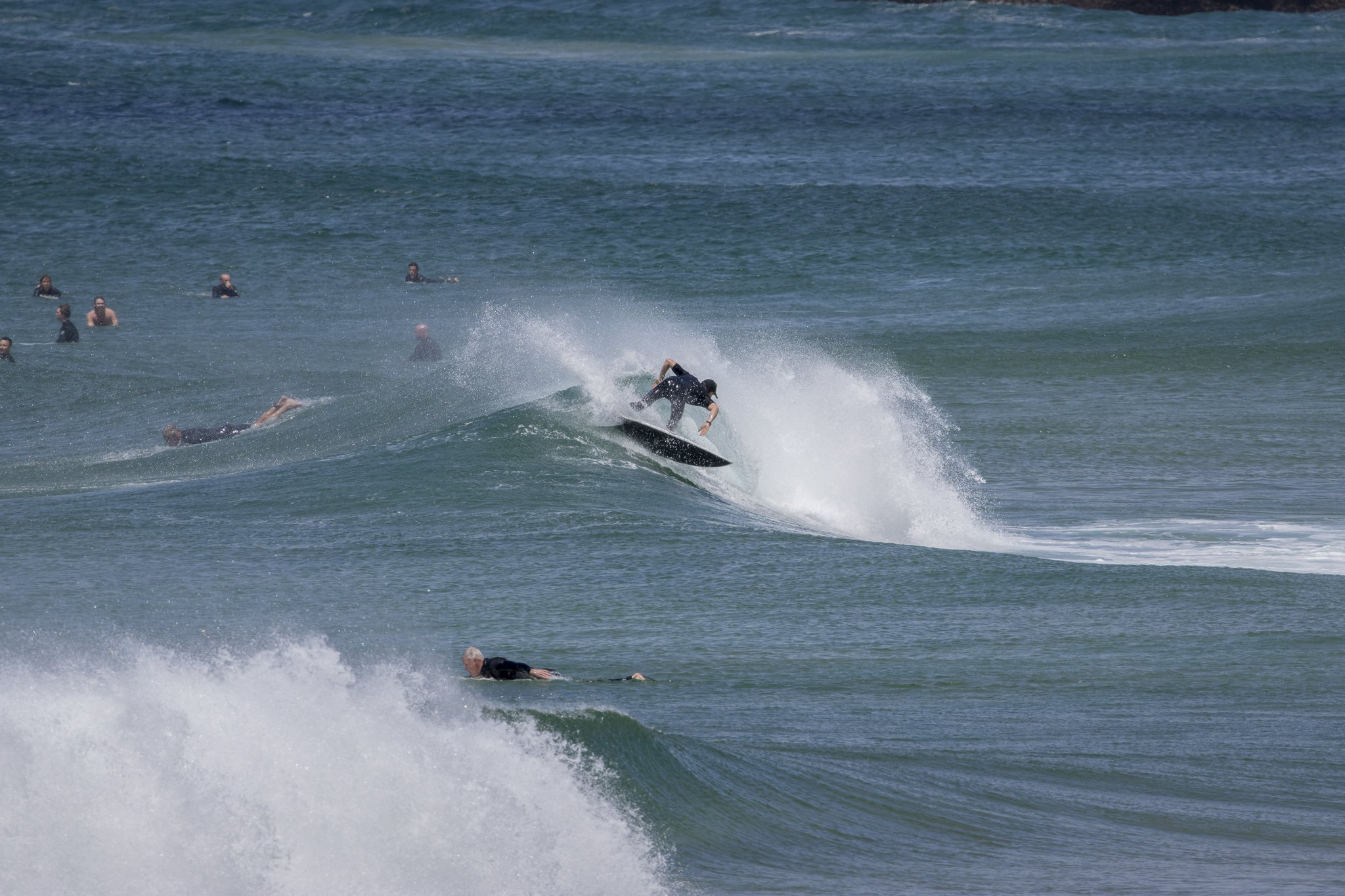 A surfer rides a wave at the beach, surrounded by other swimmers and surfers in the water.