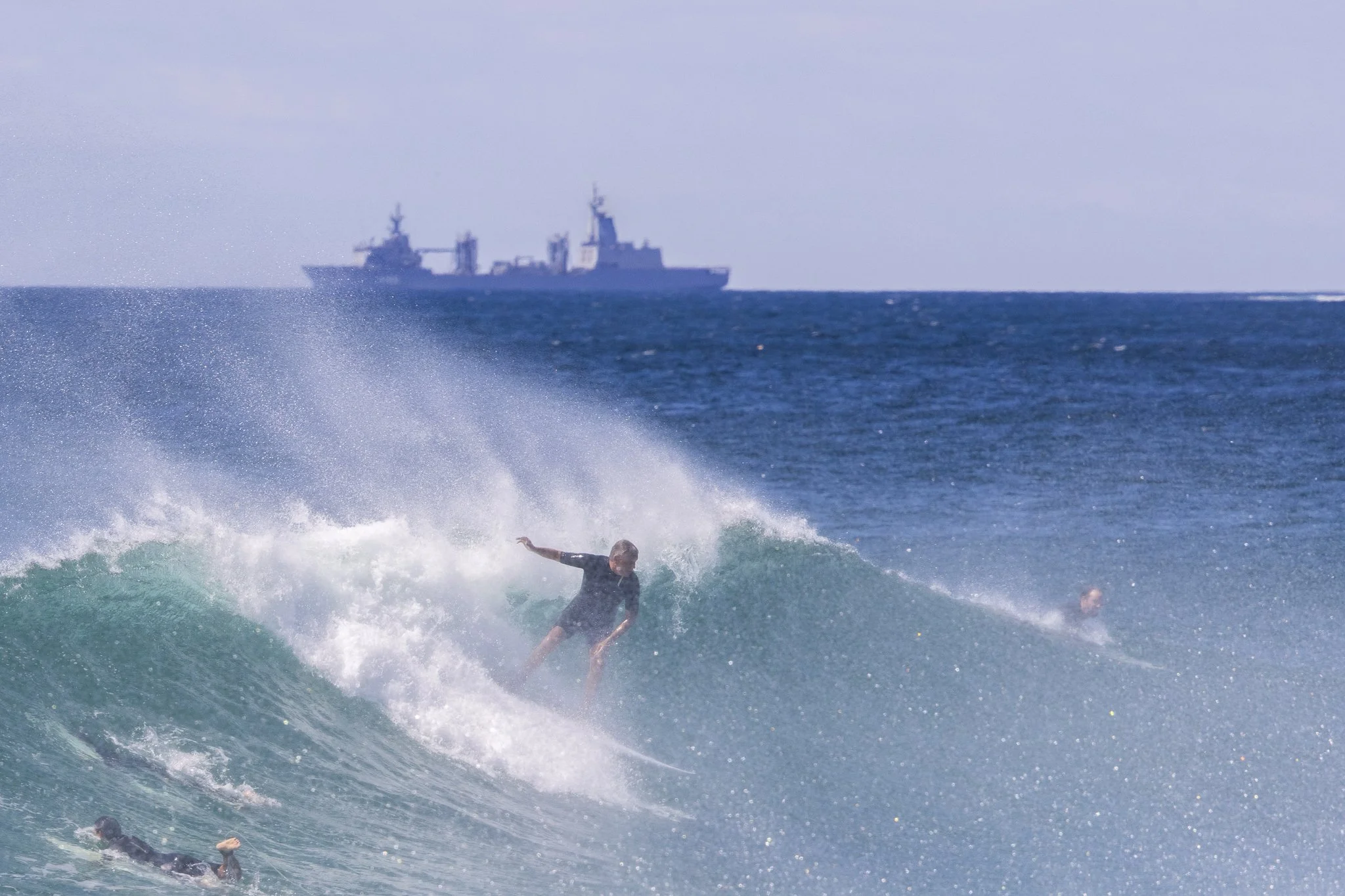 A person surfing on a wave in the ocean, with a ship in the distance.