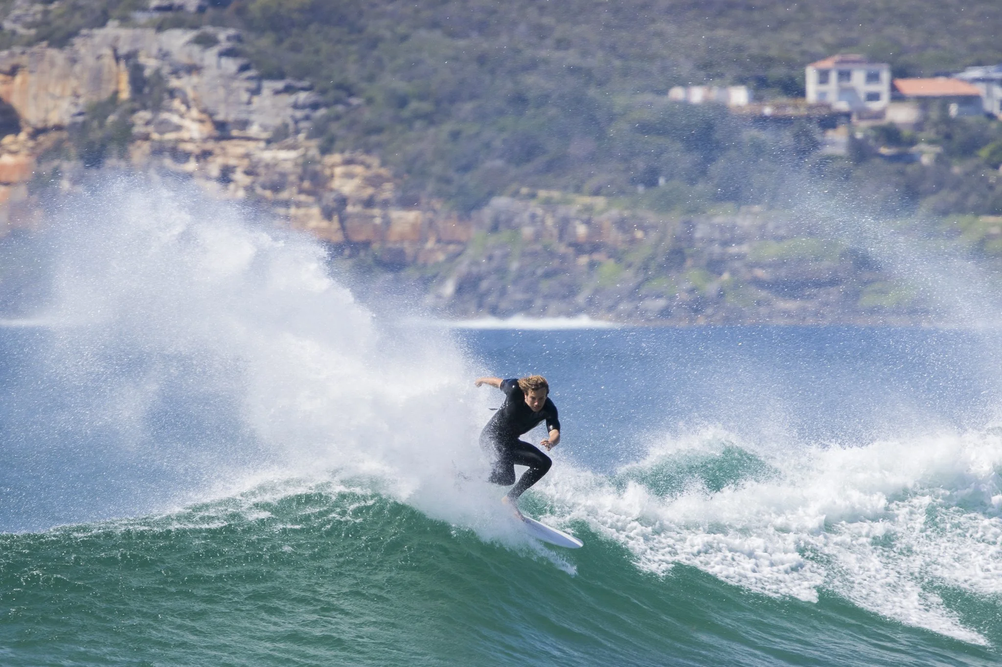 A person surfing on a wave near a rocky coastline with houses in the background.