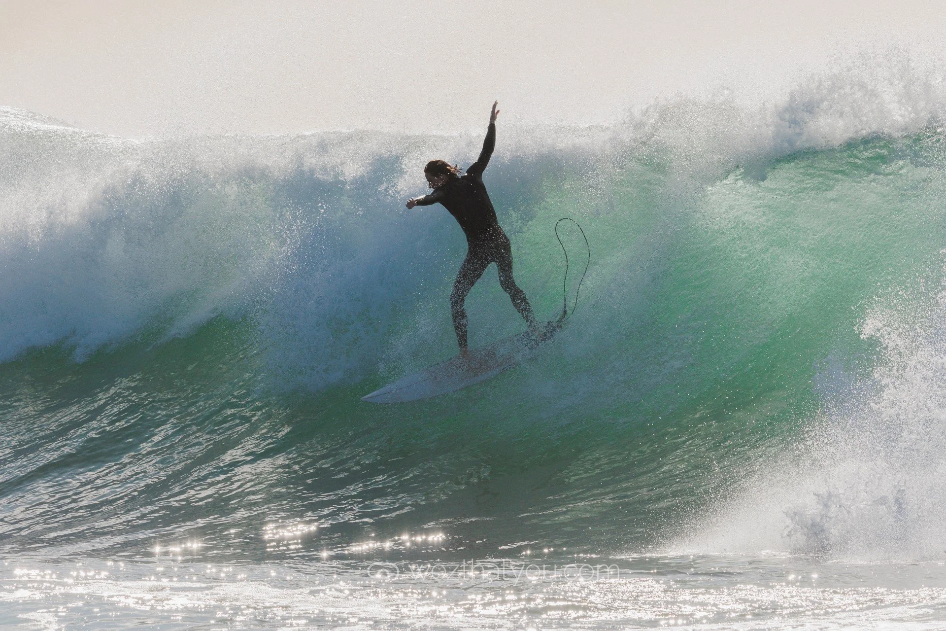 A person in black wetsuit surfing on a large wave, balancing with one arm raised and the other extended downward.