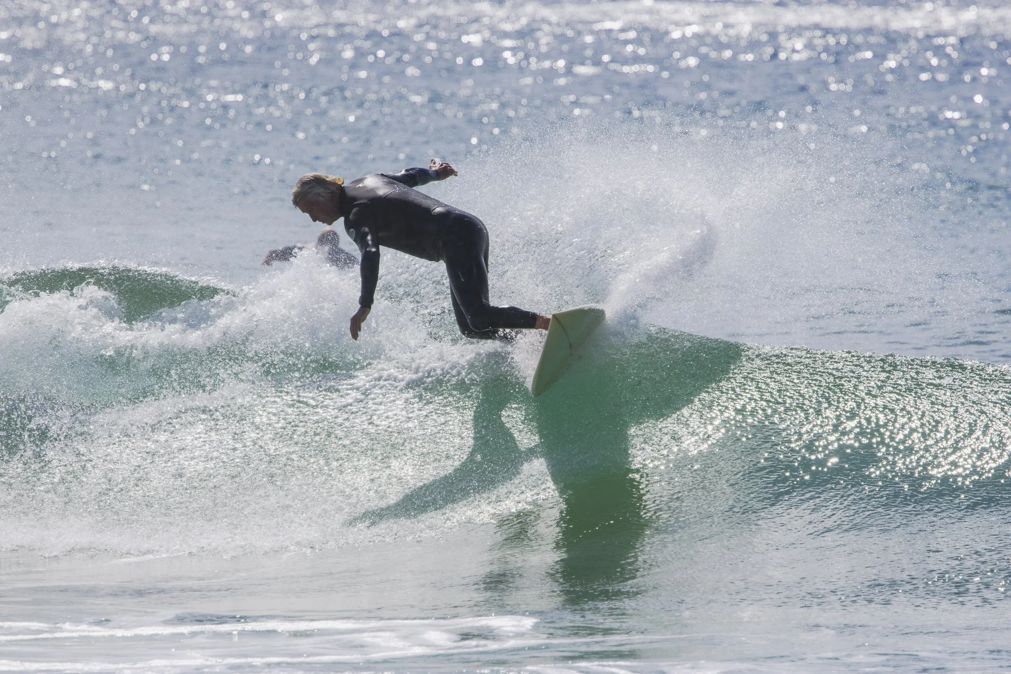 Surfer riding a wave in the ocean, wearing a black wetsuit, with sunlight reflecting on the water.