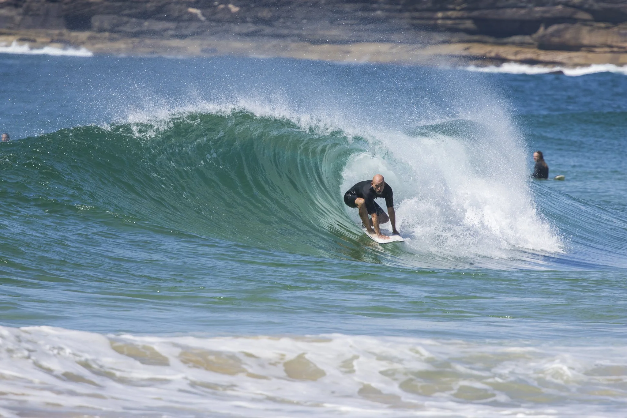 A man surfing inside the barrel of a large wave in the ocean, with a rocky shore and other surfers in the background.